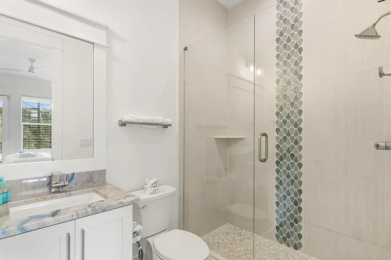 Modern bathroom featuring a white vanity with a marble countertop, a sink with a metallic faucet, a toilet with rolled towels on top, a glass-enclosed shower with pebble tile flooring, and a decorative vertical strip of gray and white pebble tiles.
