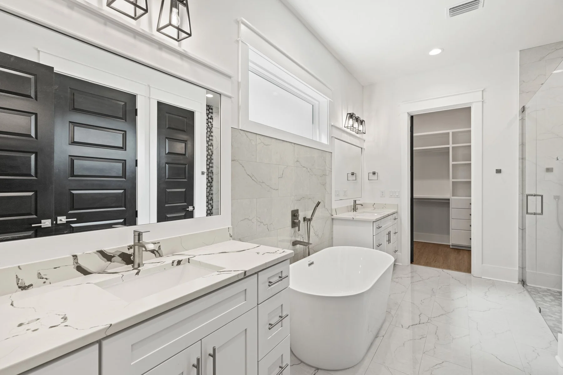 Modern bathroom with white marble countertops, a free-standing bathtub, a large mirror, white cabinetry, a walk-in closet, and black doors