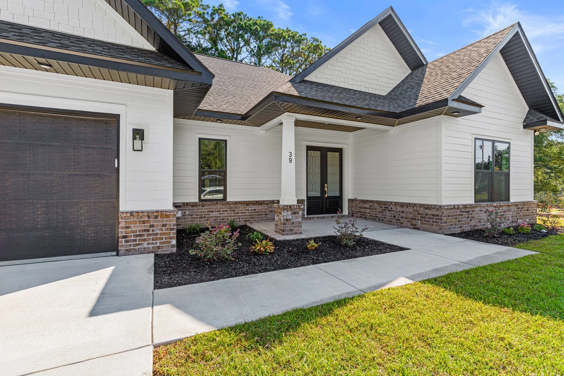 Front view of a modern house with white siding, brick accents, black door, and a garage door. The house has a peaked roof with dark shingles and a small front porch with shrubs and a concrete walkway.