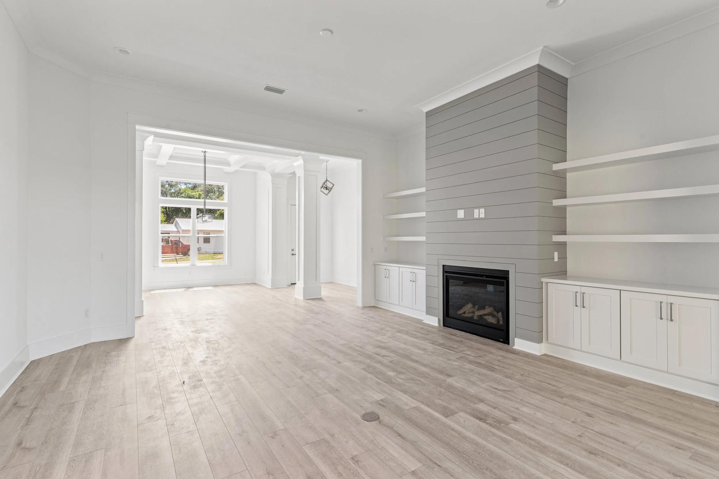 Empty modern living room with white walls, large windows, a fireplace with gray shiplap, and floating white shelves.
