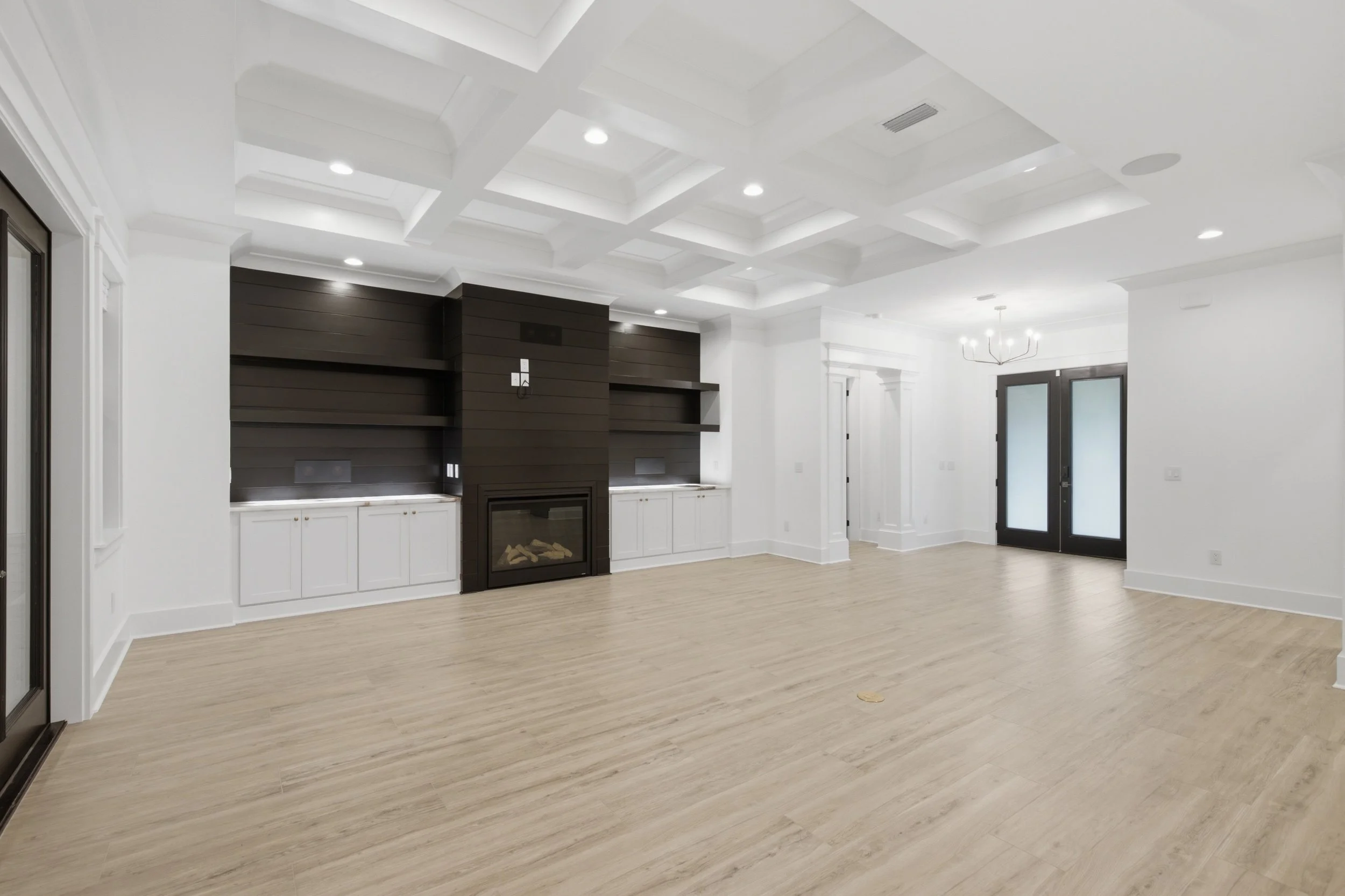 Empty living room with high tray ceiling, white walls, black double doors with glass panels, light wood flooring, built-in white cabinets, and a black fireplace with black shiplap wall behind it.
