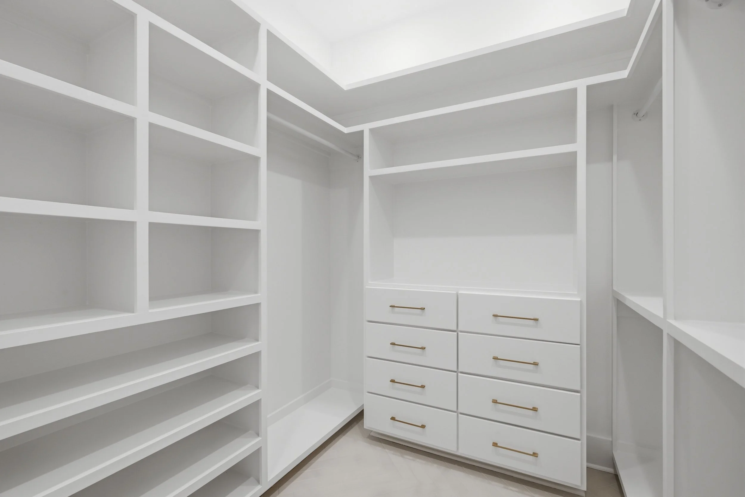 Empty white walk-in closet with built-in shelves on the left and drawers with brass handles on the right.