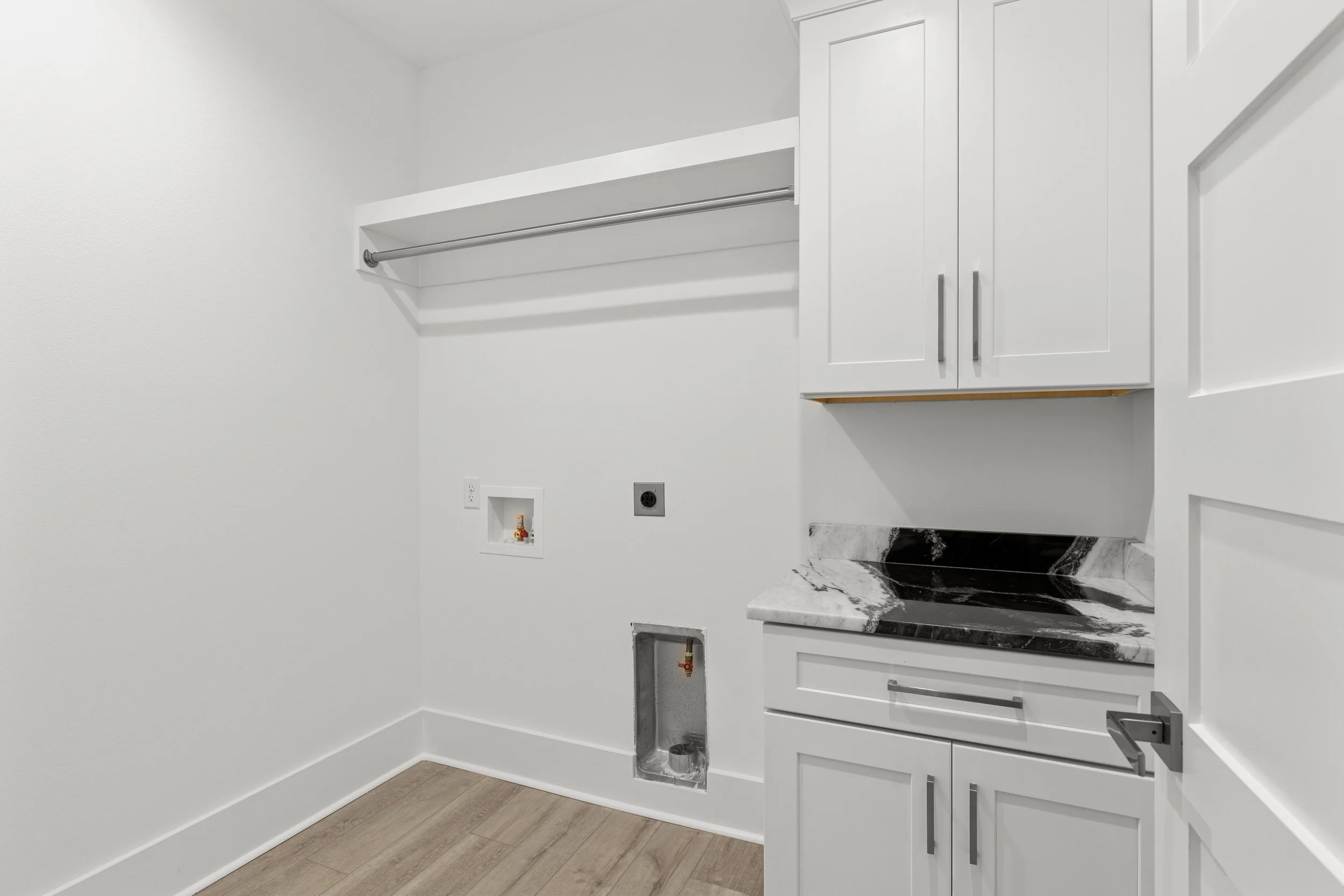 Empty laundry room with white cabinets, black and white marble countertop, and plumbing connections for appliances on a light wood floor.