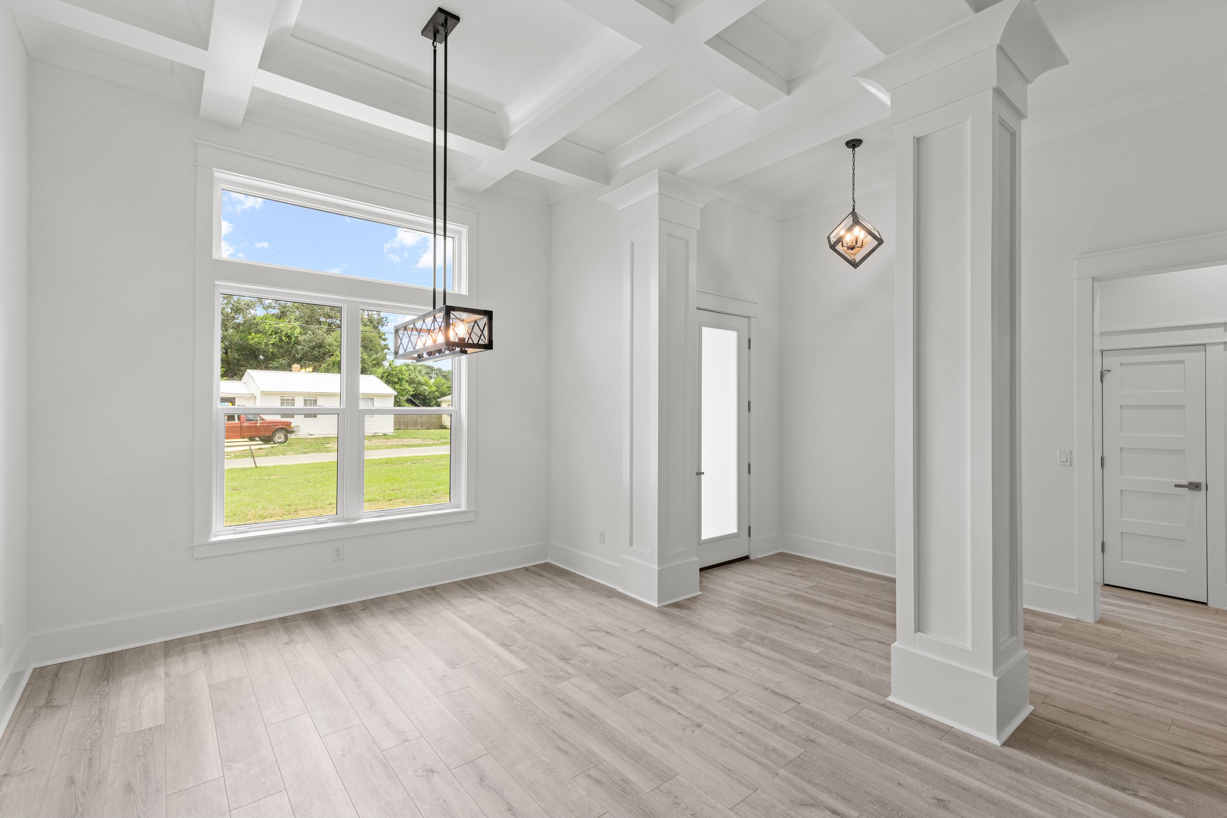 Empty white living room with high tray ceiling, large window showing a yard and house outside, modern light fixtures hanging from ceiling, light wood flooring, and partial view of a white door.