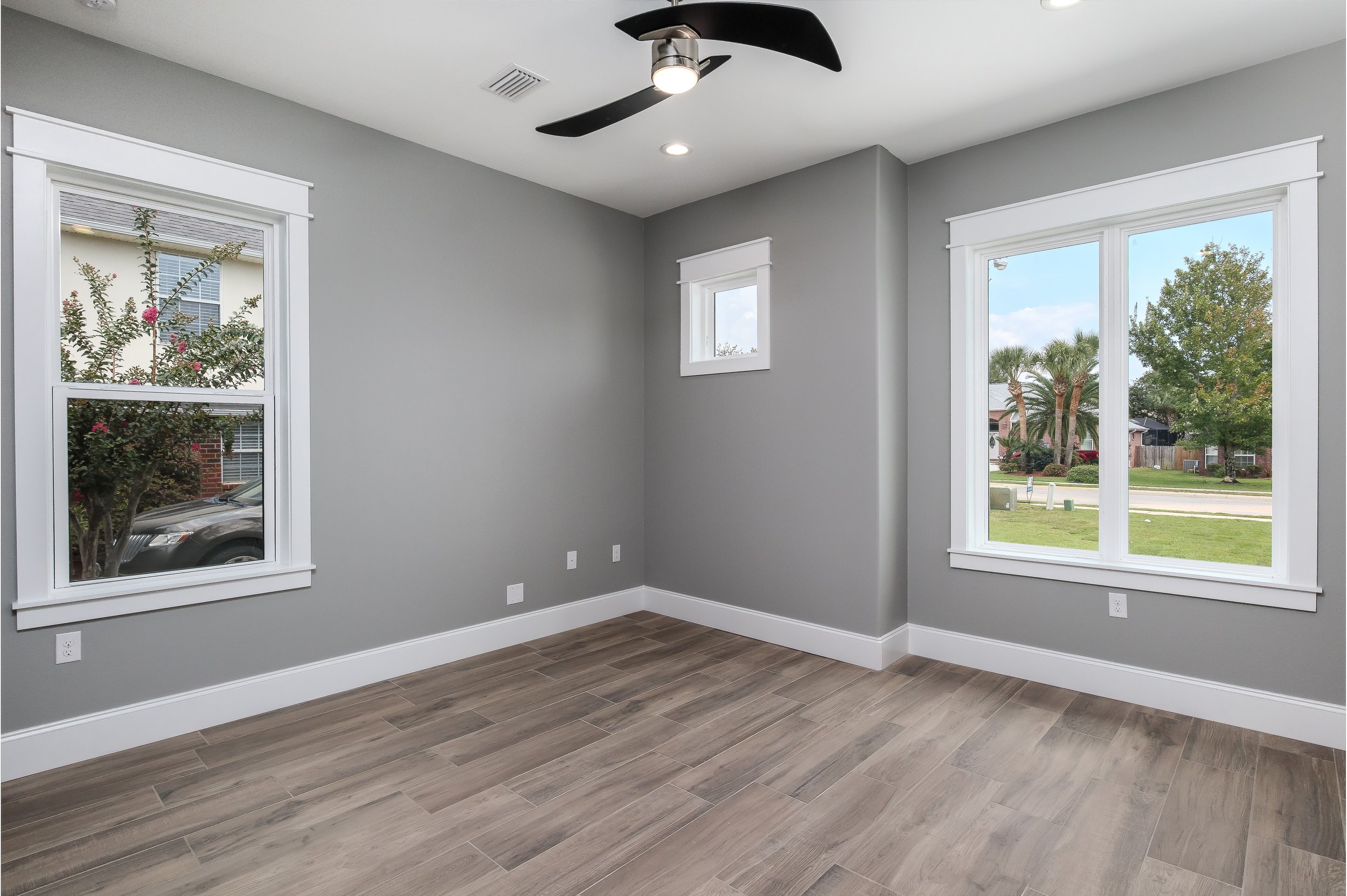 An empty room with gray painted walls, white trim, multiple windows overlooking a suburban neighborhood, and wood-look tile flooring. Ceiling fan with black blades and built-in light is installed on the ceiling.