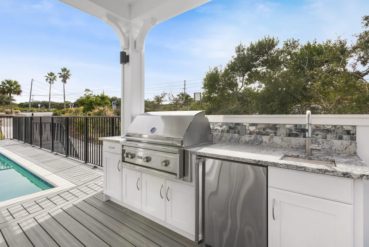 Outdoor kitchen with stainless steel grill, sink, and granite countertop on a patio with a swimming pool and trees in the background.