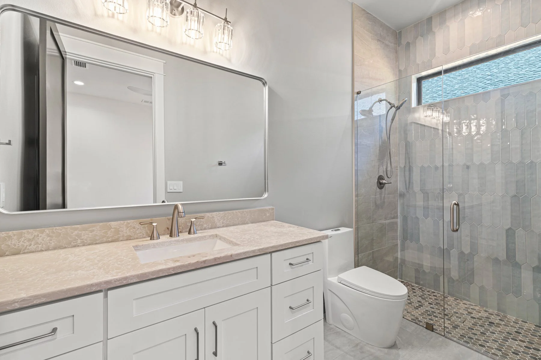 Modern bathroom with a white vanity, marble countertop, large mirror, and a glass-enclosed shower with gray hexagonal tiles and a small horizontal window.