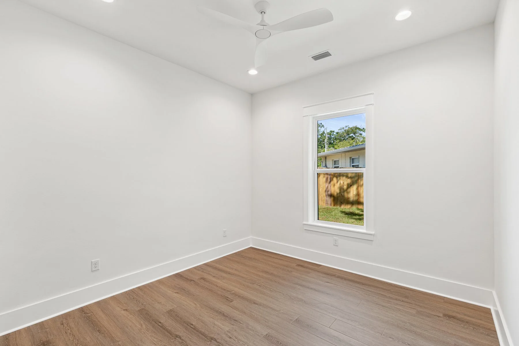 Empty room with white walls, a window showing a view of a backyard with a fence and house, a ceiling fan, and wood flooring.