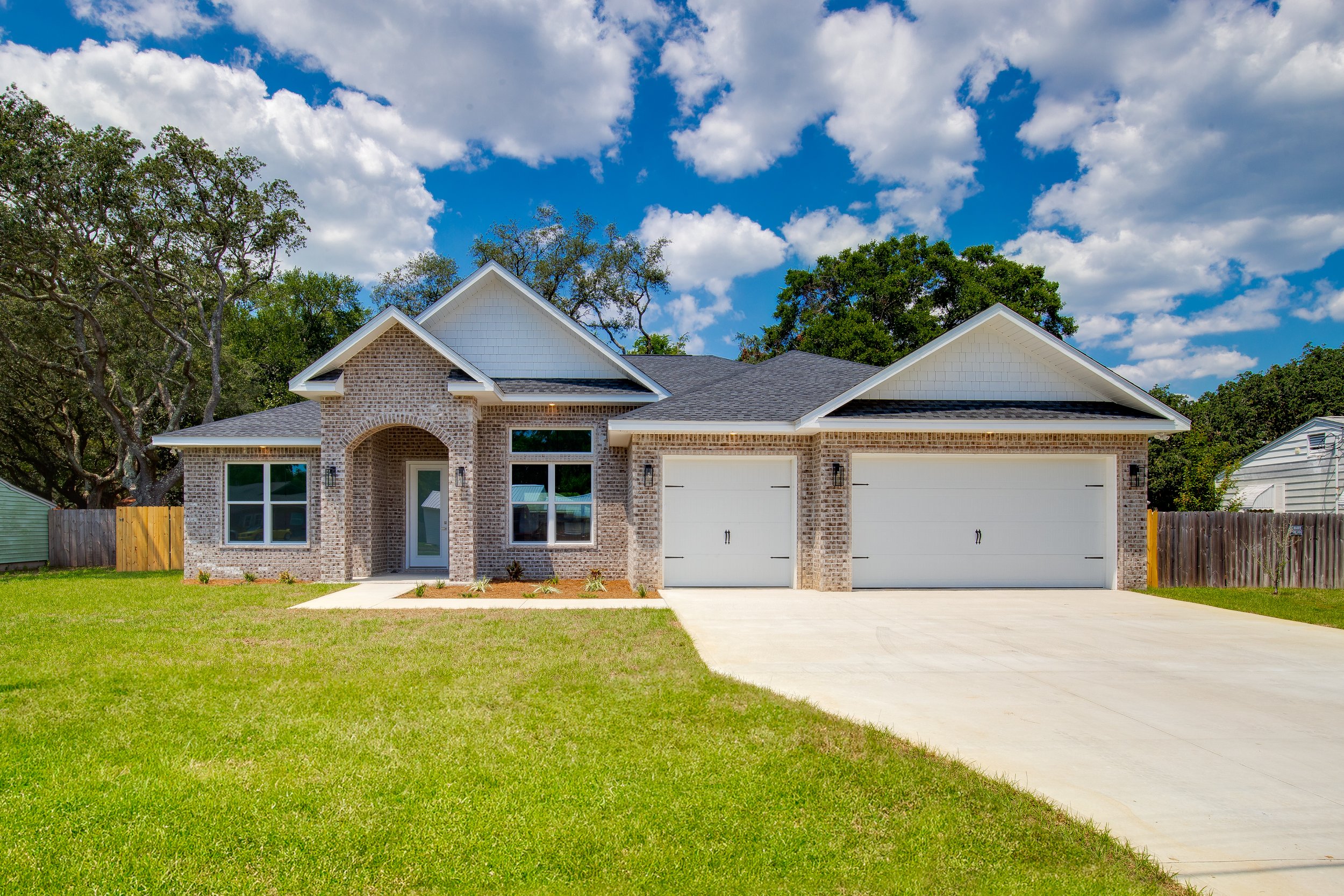 Brick house with white garage doors and a concrete driveway, green lawn, surrounded by trees, under a partly cloudy sky.