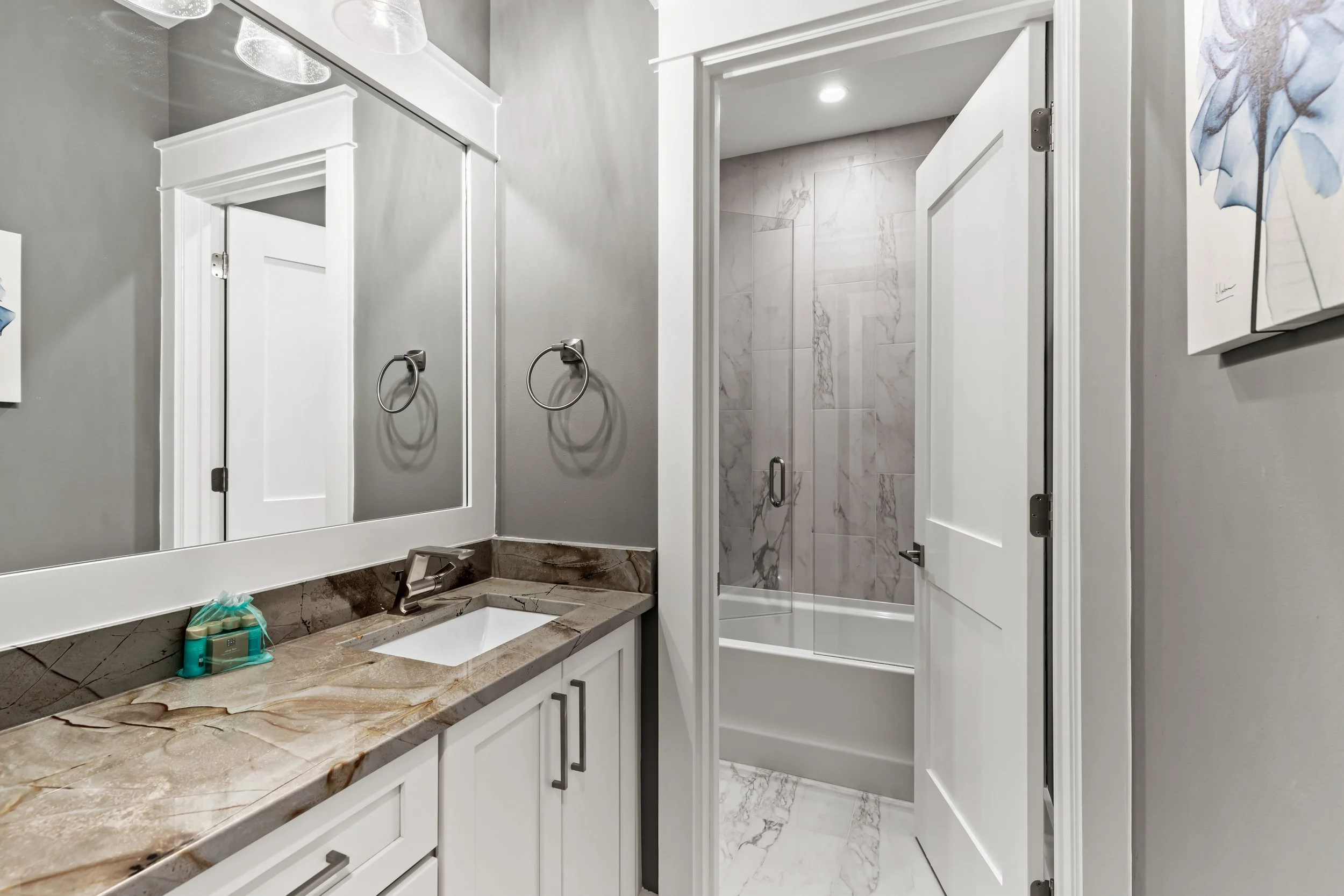 A bathroom with a gray wall, a large mirror, and a marble countertop with a sink. There is a white cabinet with dark handles below the counter and a soap dispenser on the counter. The bathroom has a shower with glass doors and marble tiles. There is a white door with trim and a silver handle leading to the shower. A piece of blue artwork is partially visible on the right side of the image.
