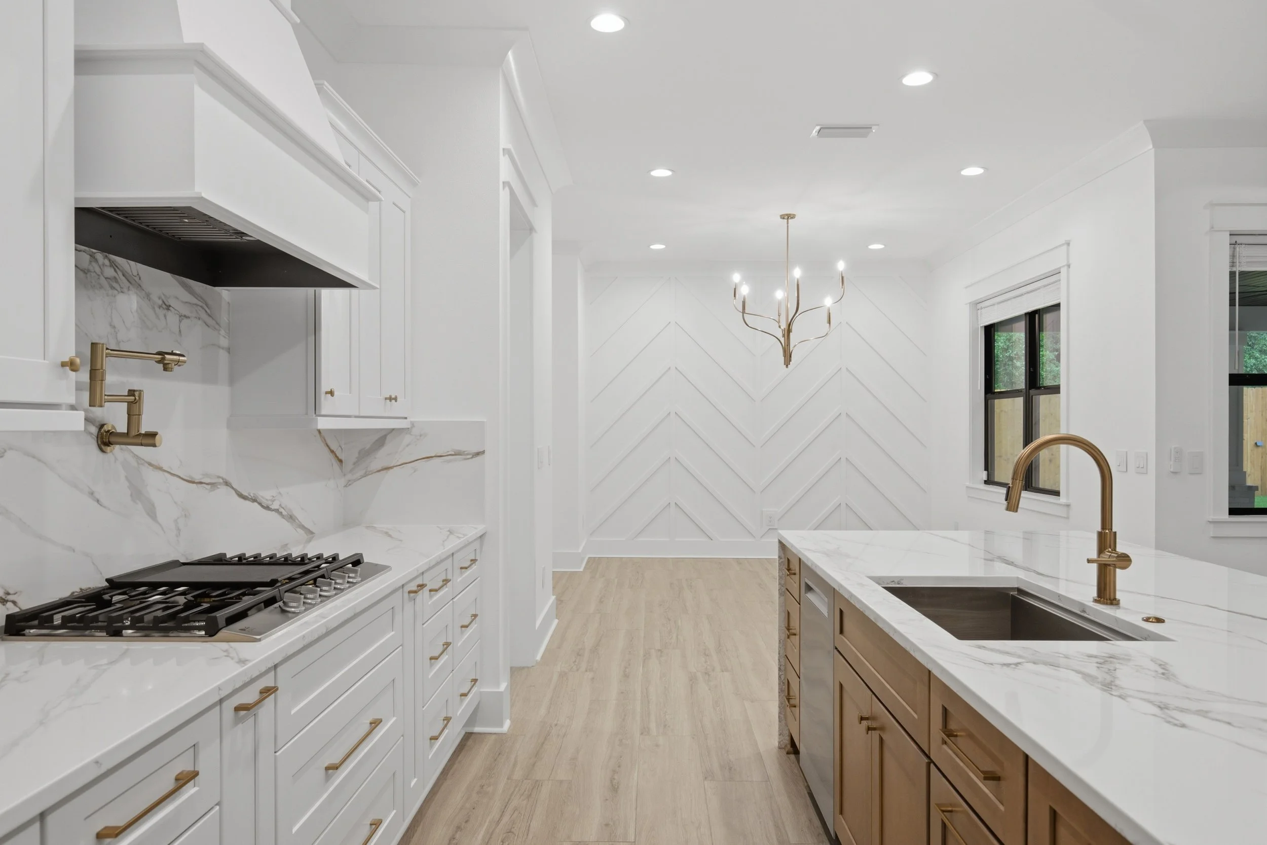 Modern kitchen with white cabinets, marble countertops, and gold fixtures. There is a chandelier hanging from the ceiling, a window with view outside, and a gas stove on the left.