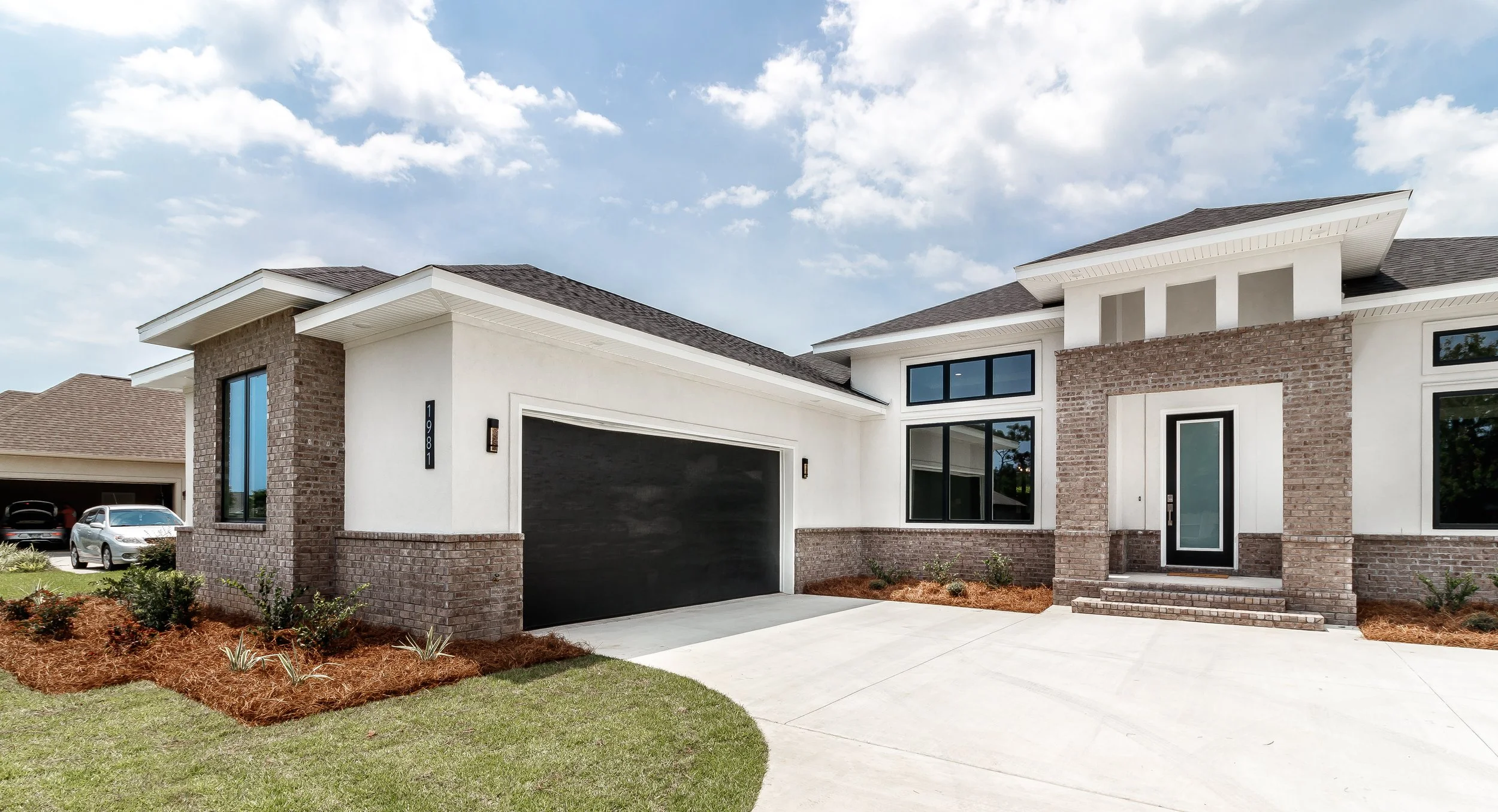 Modern two-story house with white walls, brick accents, large windows, and a black garage door. Front yard with mulch, small plants, concrete driveway, and stairs leading to the front door.