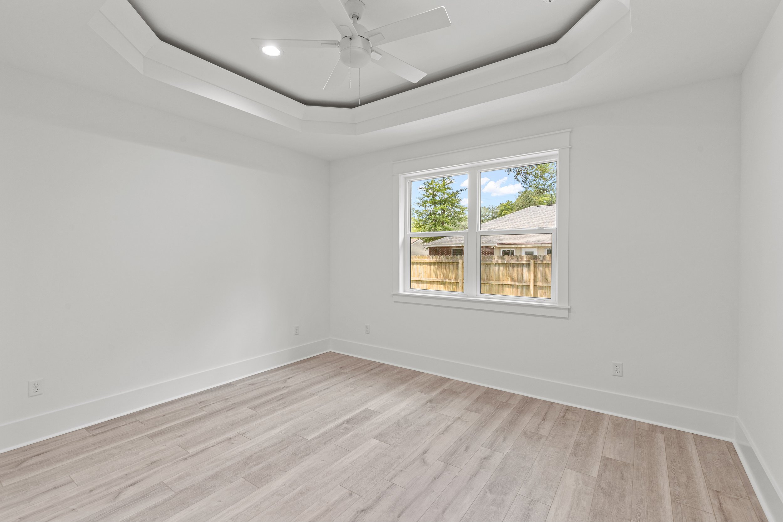 Empty room with white walls, light wood flooring, a large window showing a backyard with a wooden fence and trees, a ceiling fan, and recessed lighting.