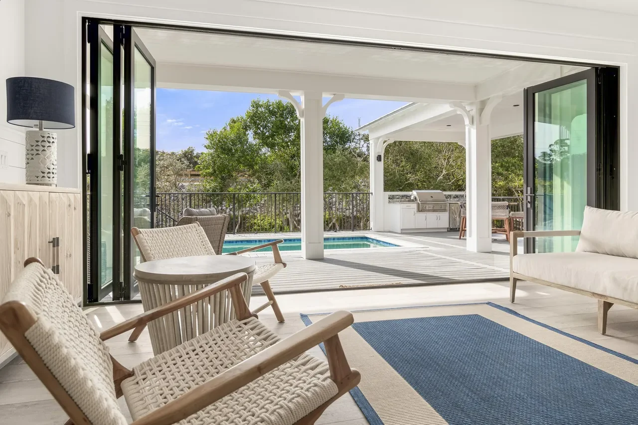 Indoor living room opening to outdoor pool area with chairs, sofa, and grill.