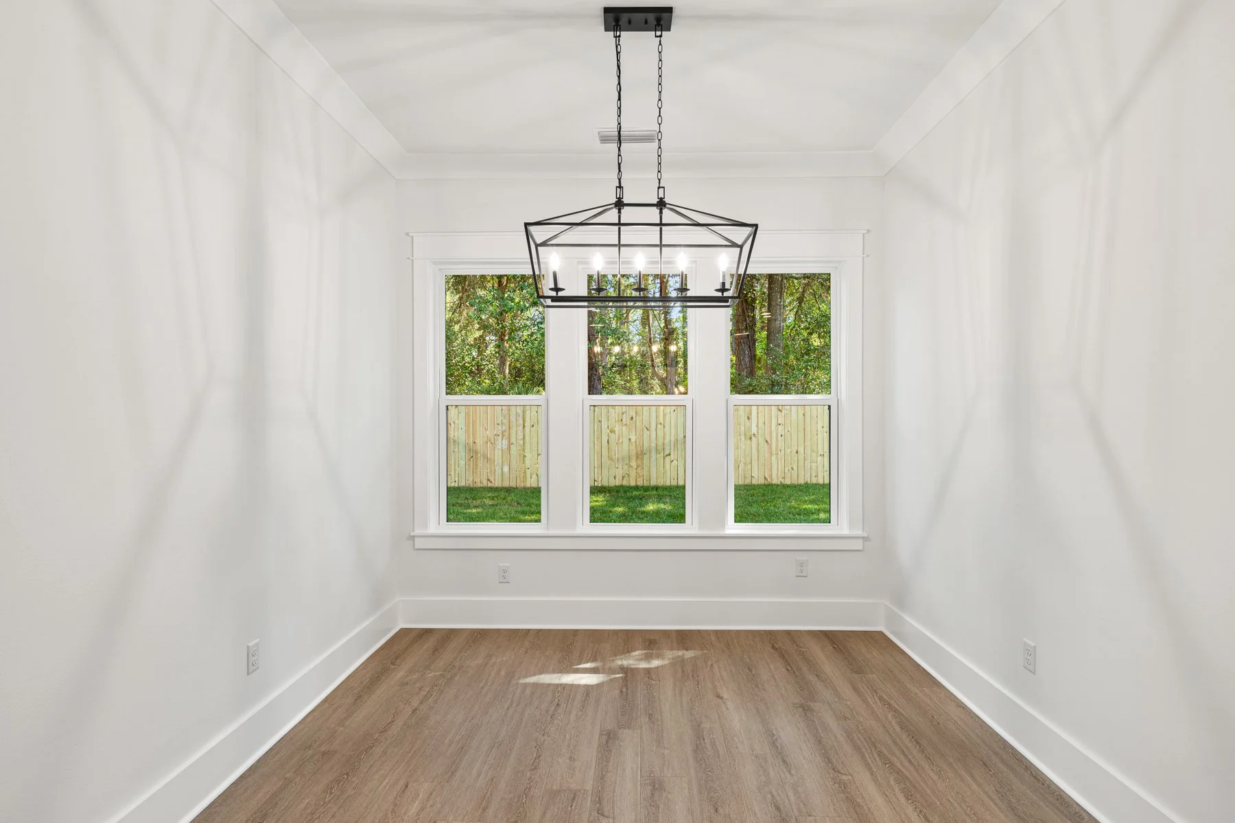 Empty room with white walls, large window with three sections showing a backyard with trees and a wooden fence, and a black geometric chandelier with six candle-like lights hanging from the ceiling.