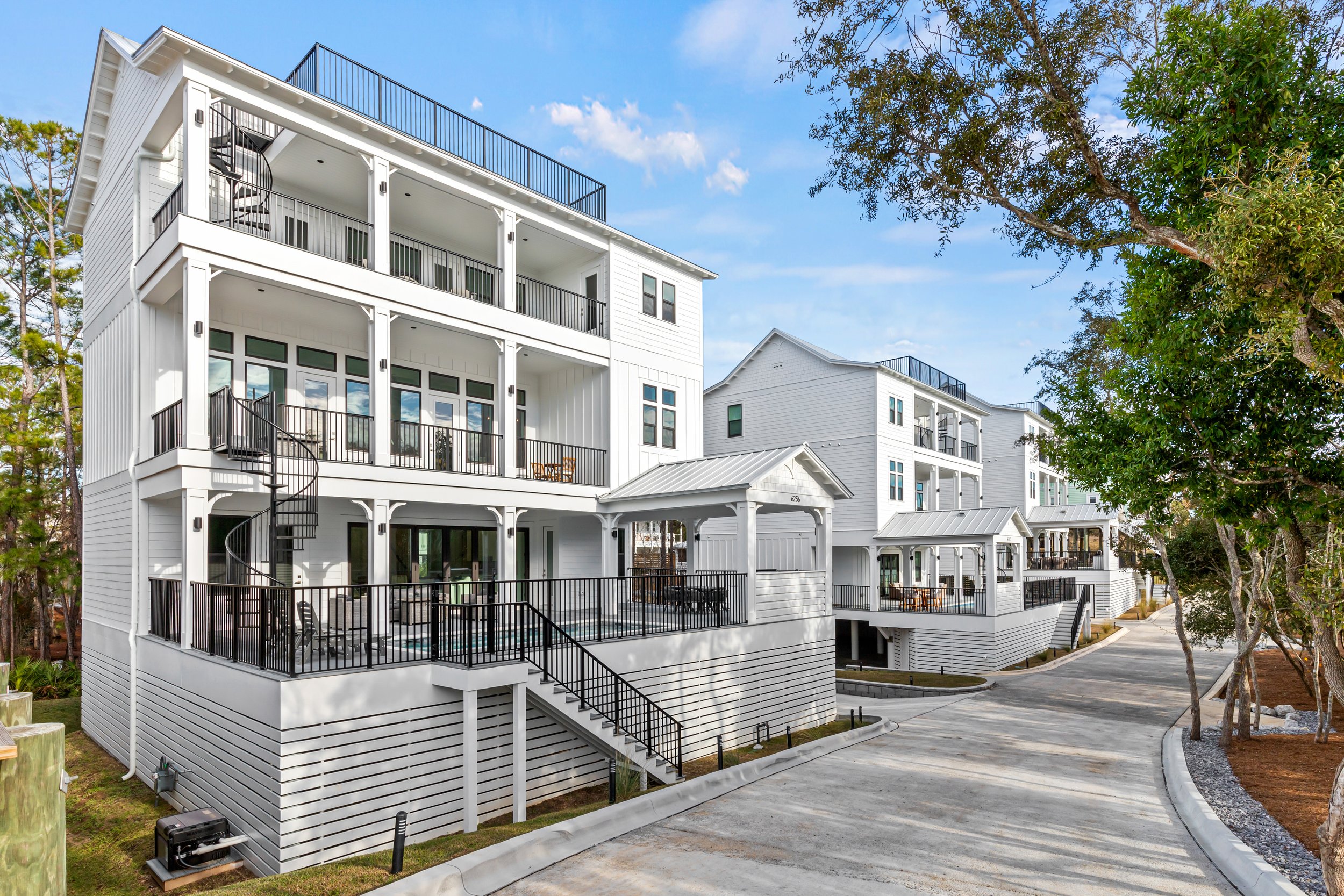 White multi-story residential buildings with black railings on balconies, spiral staircases, large windows, surrounded by trees and a paved driveway, under a partly cloudy sky.