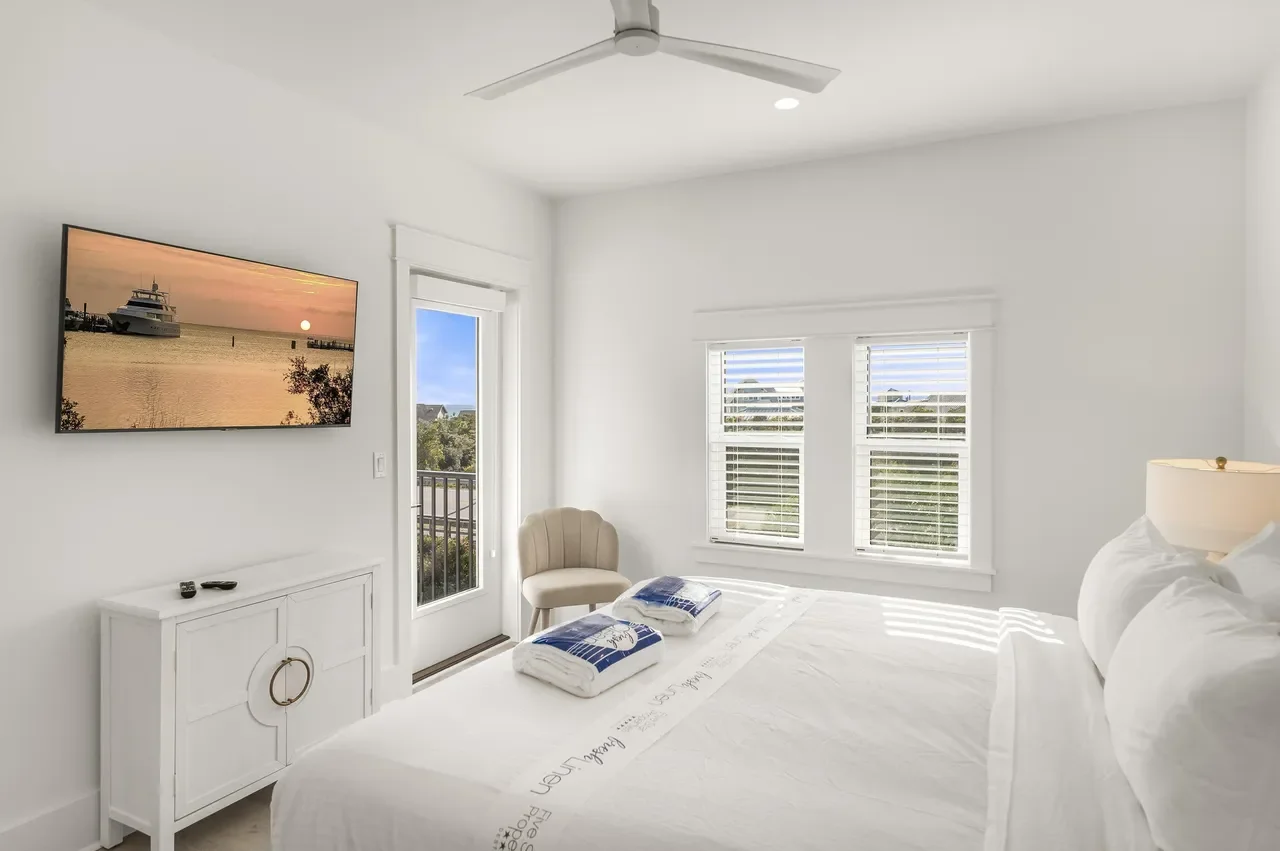 Bright bedroom with white walls, window blinds, and a bed with white bedding. A wall-mounted TV shows a boat at sunset. There's a small beige chair, a white cabinet, and a ceiling fan.