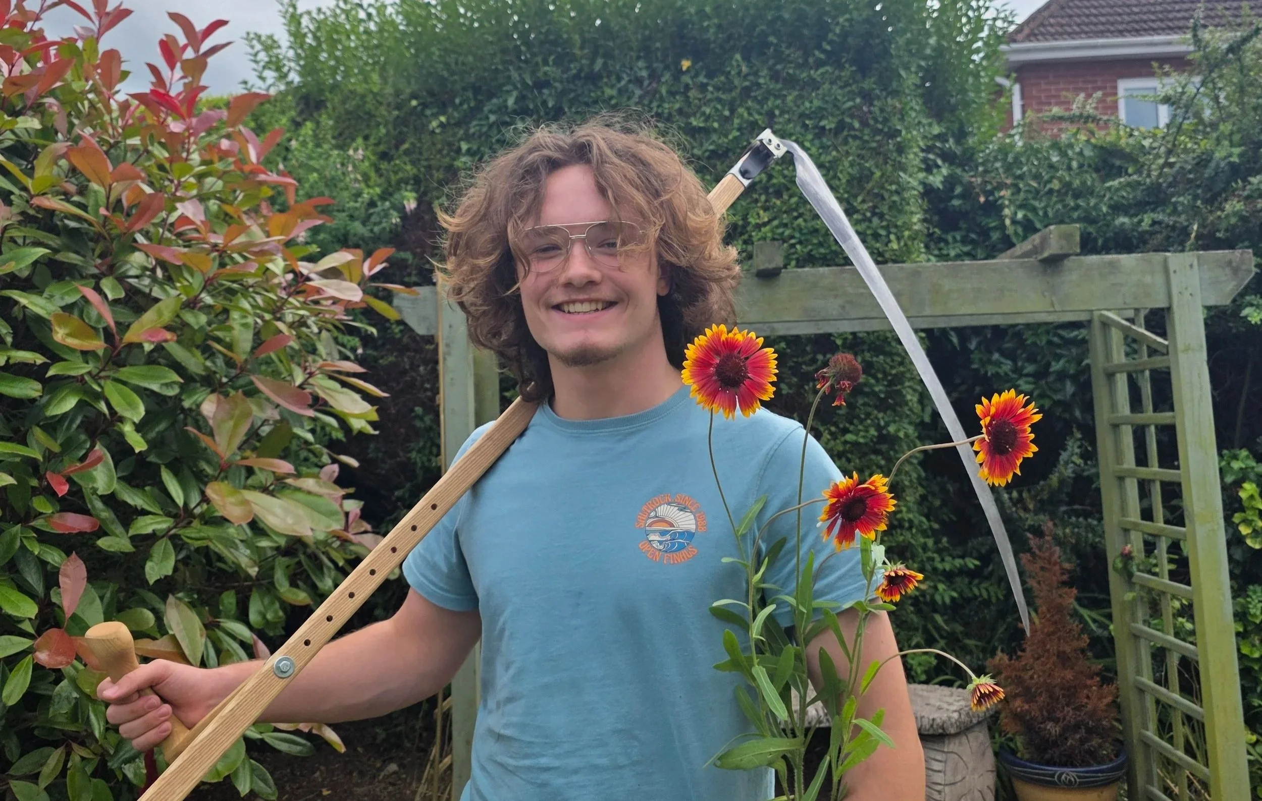 A young man with curly hair, wearing glasses and a blue t-shirt, holding a bouquet of orange and yellow flowers in a garden with green plants and a wooden structure in the background.
