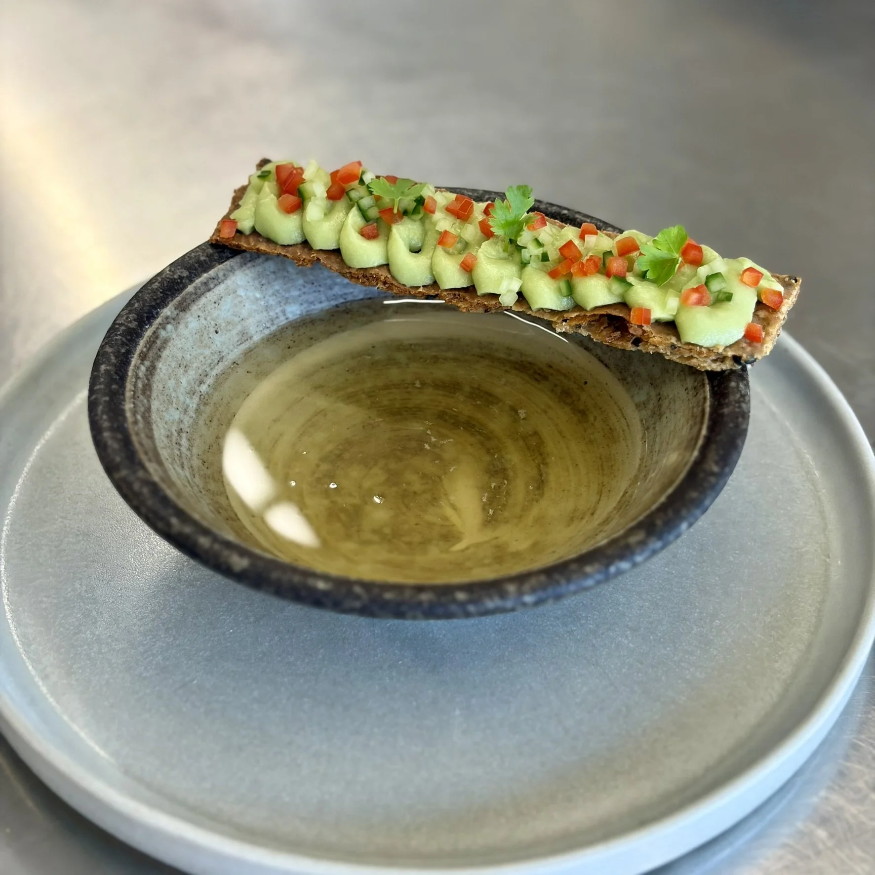Close-up of a dish with creamy guacamole topped with chopped red and green peppers and cilantro, served on a crispy tortilla chip over a bowl of melted cheese or sauce in a ceramic bowl.