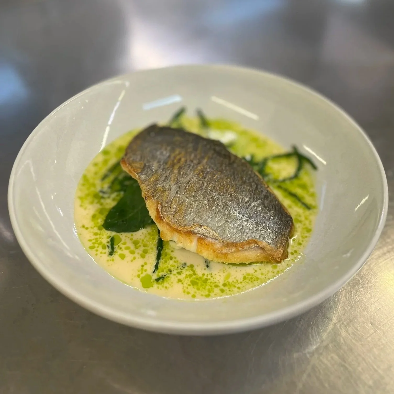 Pan-seared fish fillet with crispy skin in a creamy green sauce, garnished with fresh herbs, served in a white bowl.