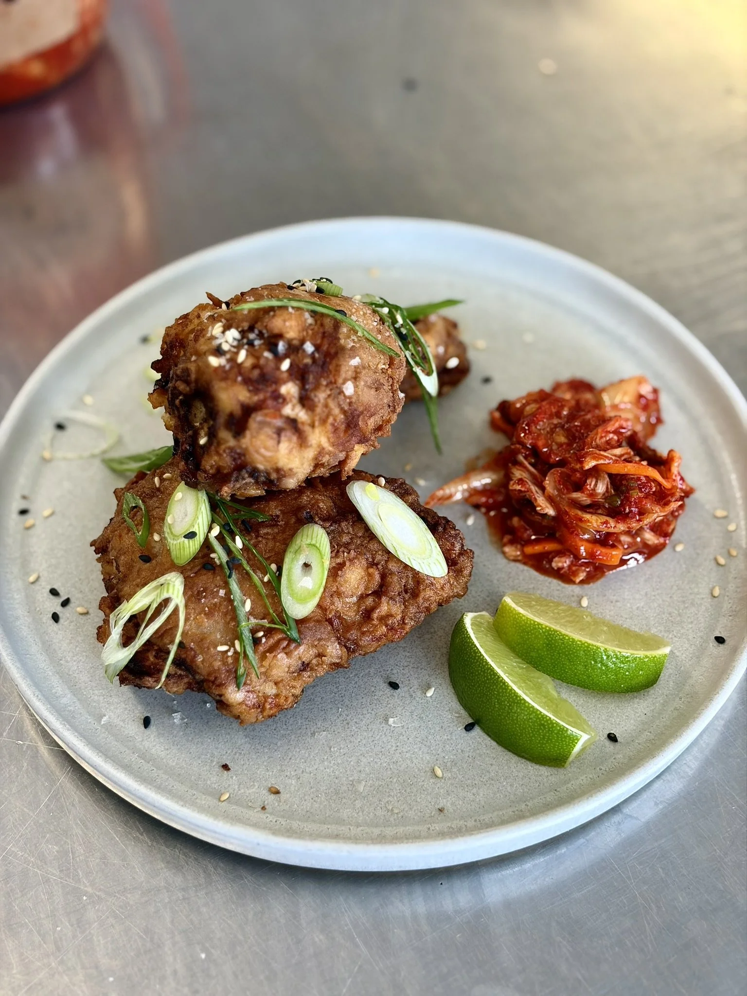 Fried chicken drumsticks garnished with sliced green onions and sesame seeds, served with lime wedges and kimchi on a gray plate.