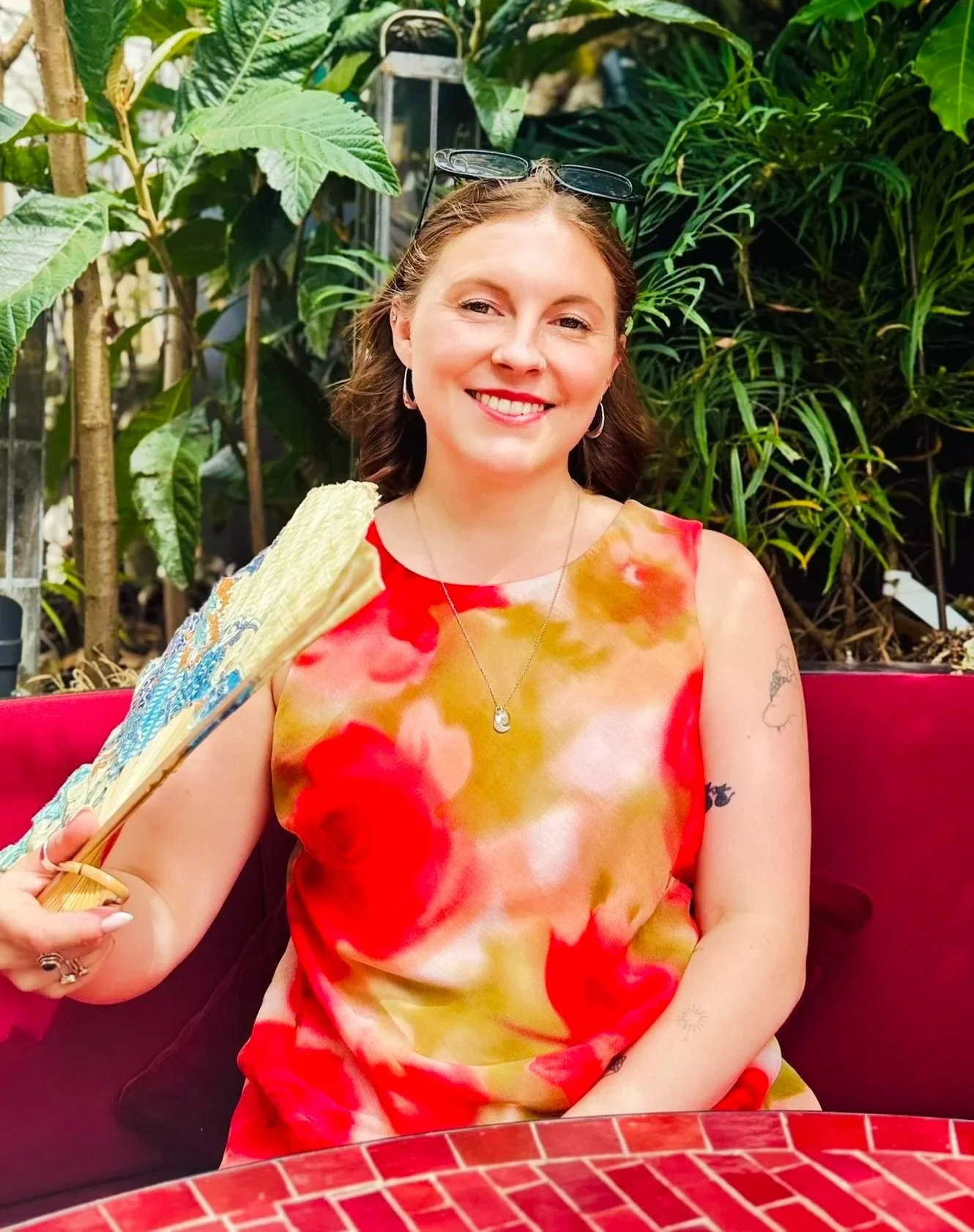 Woman with short brown hair wearing a colorful floral dress, holding a fan, sitting at a table with a red tiled surface in an outdoor setting with green plants in the background.