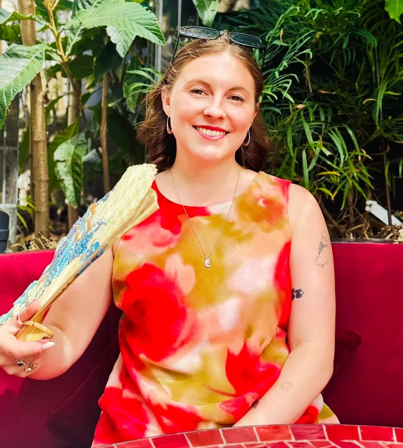 A woman with shoulder-length brown hair smiling, wearing a colorful sleeveless dress with red, yellow, and pink patterns, holding a fan, sitting outdoors in front of green plants.