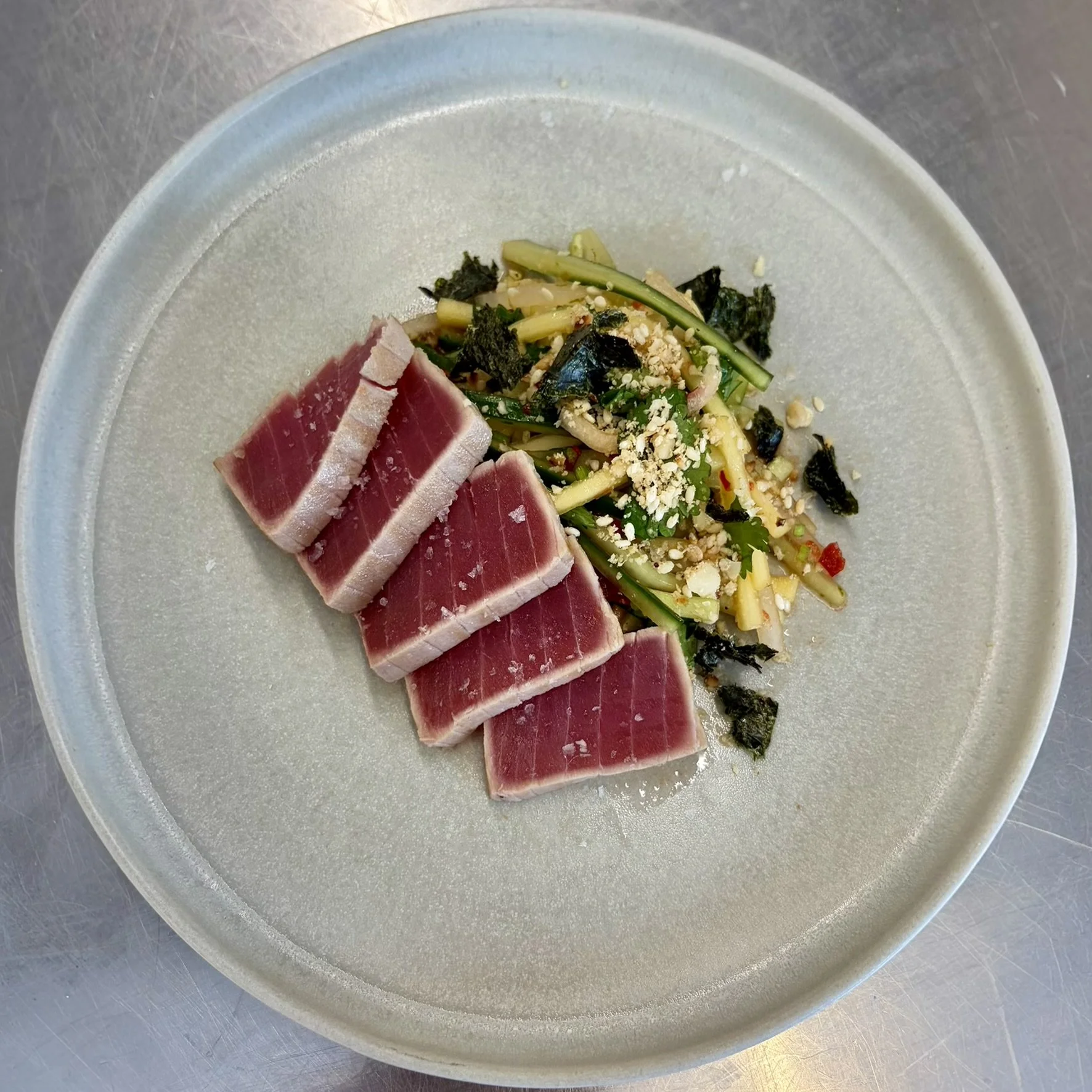 Sliced rare tuna served with a side of vegetable salad garnished with crushed nuts on a white disposable plate.