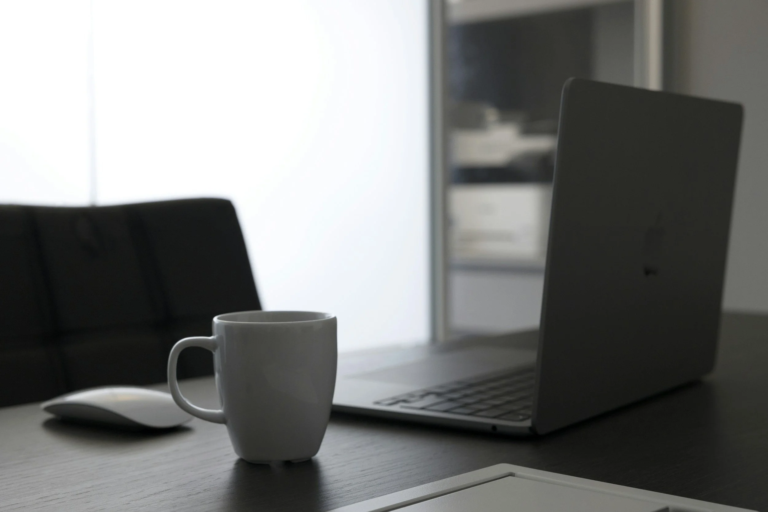 An office desk with a computer, a white coffee mug, and a white computer mouse in the foreground. A black chair is positioned next to the desk. In the background, there is a shelving unit with various items, and a bright window providing natural light to the workspace.