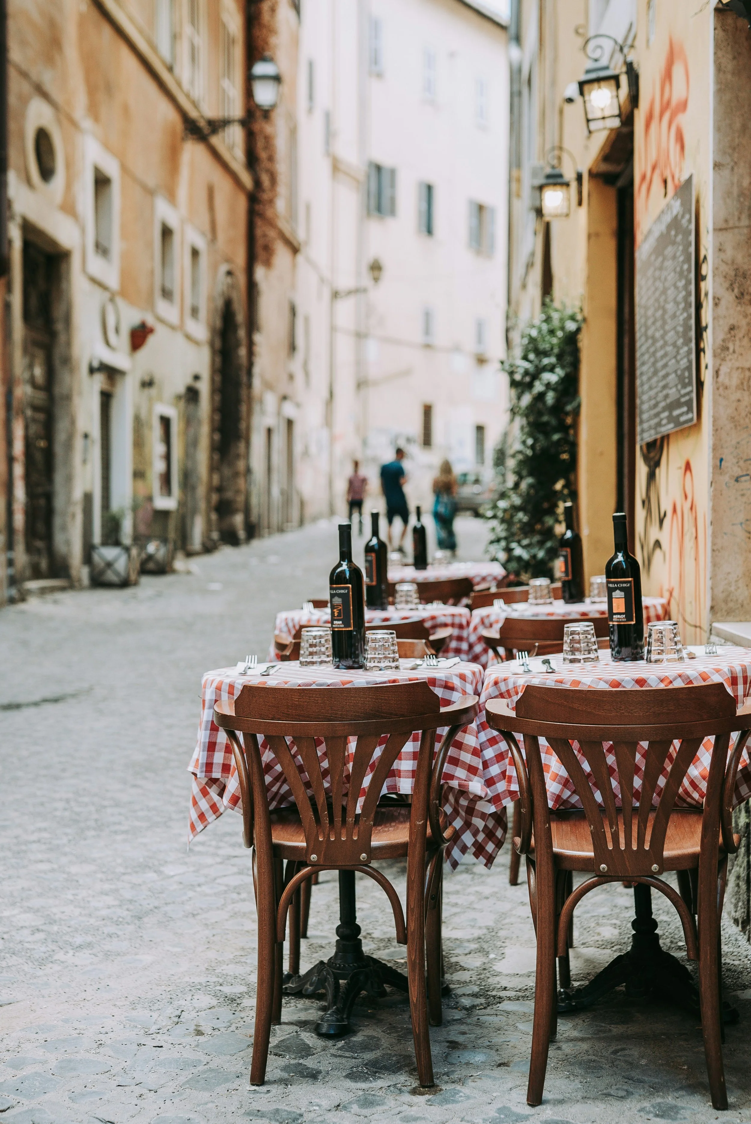 Outdoor restaurant seating with tables covered in red and white checkered tablecloths, wine bottles, and glasses, set on a cobblestone street in a European town, with blurred buildings and pedestrians in the background.