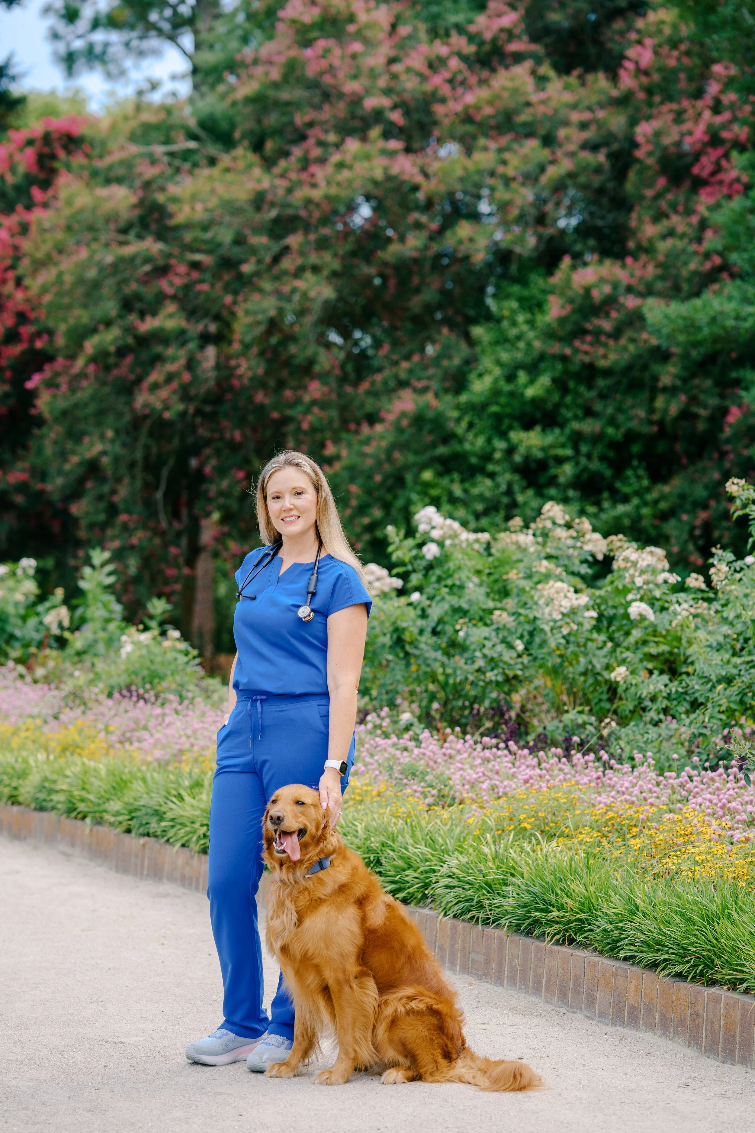 A woman dressed in blue medical scrubs standing in a garden with a Labrador retriever dog. The woman has blonde hair, is smiling, and wears a smartwatch. The dog is sitting, has a shiny golden coat, and looks happy with its tongue out. The garden background includes pink, white, and yellow flowers, and trees with green foliage.