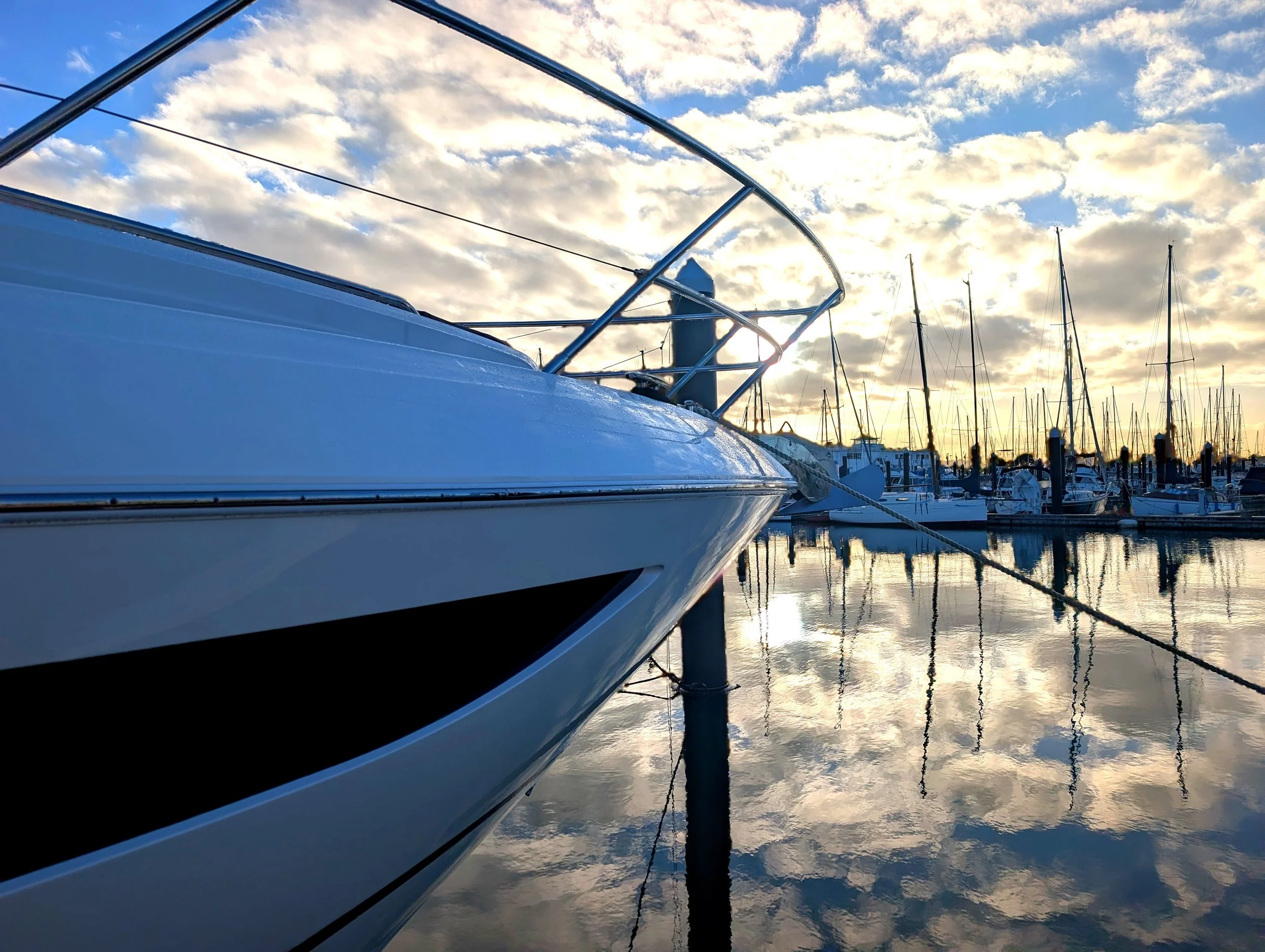 Close-up of a white yacht's deck and railing at a marina with multiple sailboats moored, cloudy sky at sunset or sunrise reflected on the calm water.
