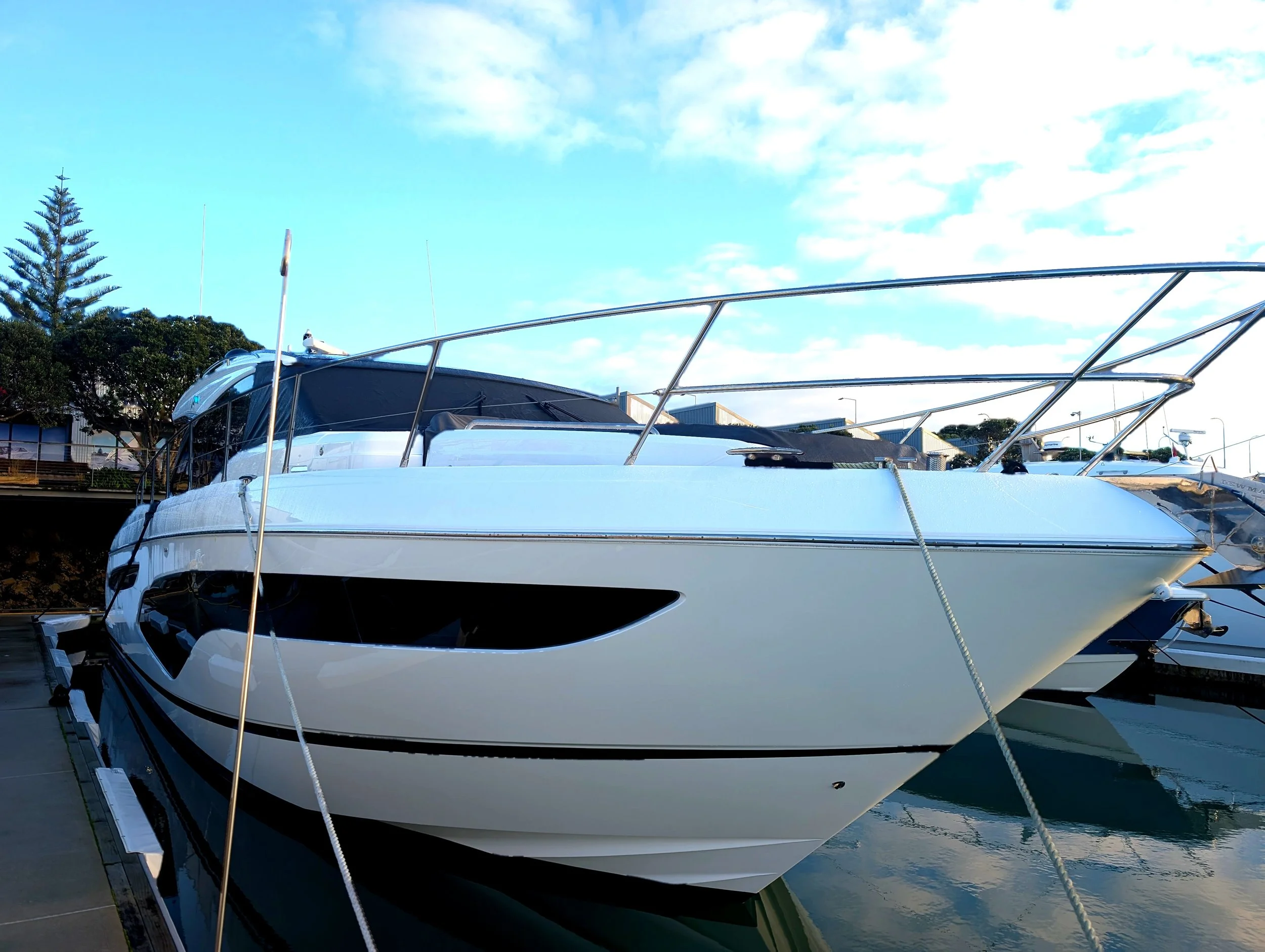 A white yacht docked at a marina, with a clear blue sky and some clouds above, and trees and buildings in the background.