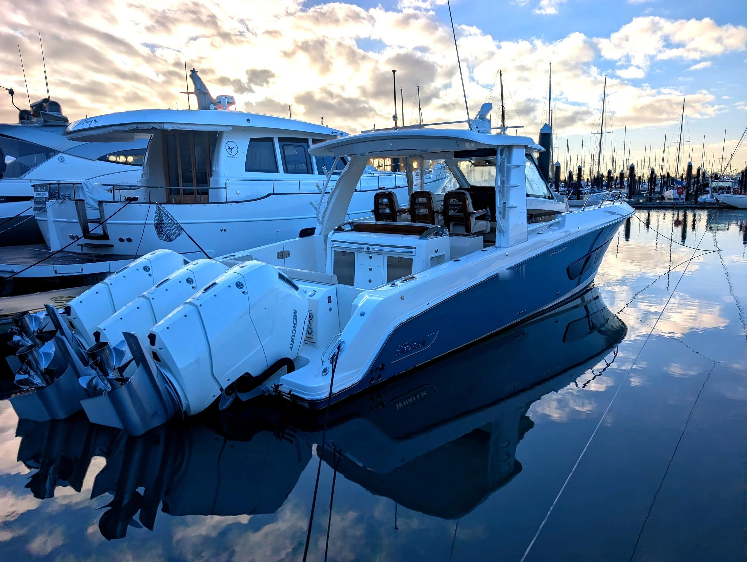 A marina with multiple boats docked on calm water during sunset. The sky is partly cloudy with sunlight breaking through, reflecting on the water's surface.