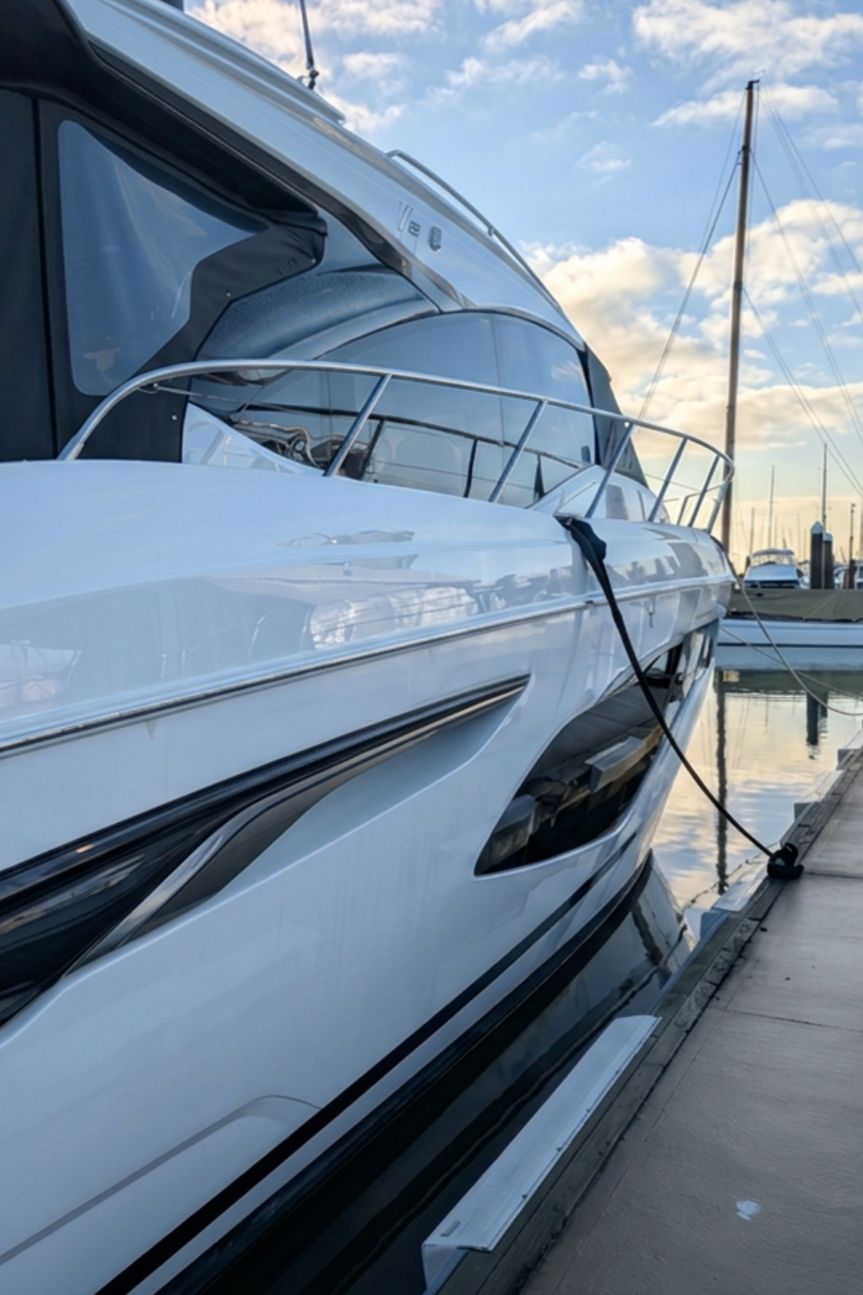 A close-up of a modern yacht docked at a marina, with calm water and a partly cloudy sky in the background.