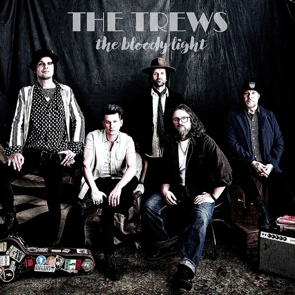 A music band called The Trews posing for a photo with five members against a dark backdrop with their band name and album title 'The Bloody Light' displayed.
