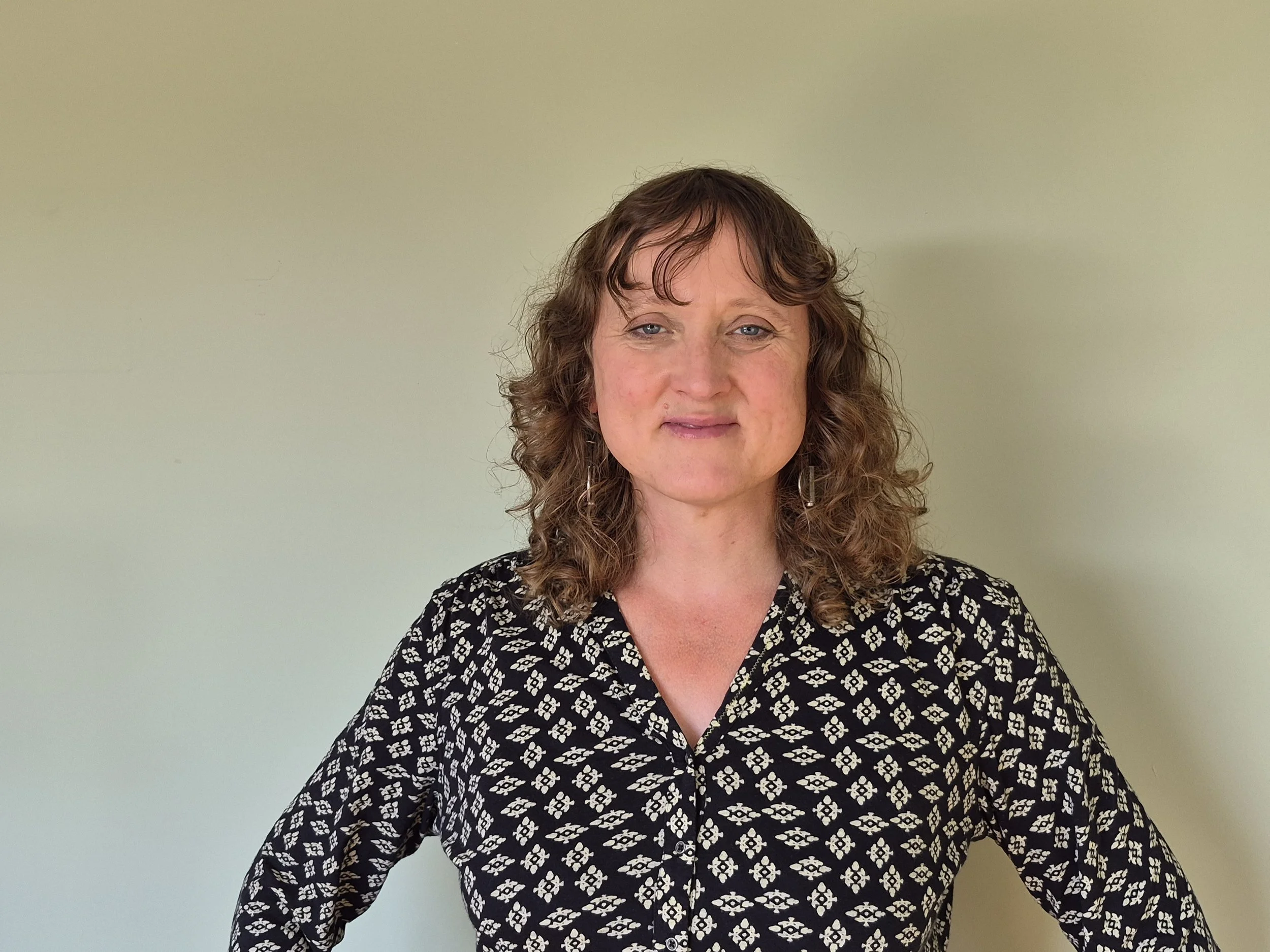 Jenny Wellwood is a white woman, 46 years old, has curly brown hair and is wearing a black and white patterned top, standing in front of a plain light green wall. She has a gentle smile and warm eyes.