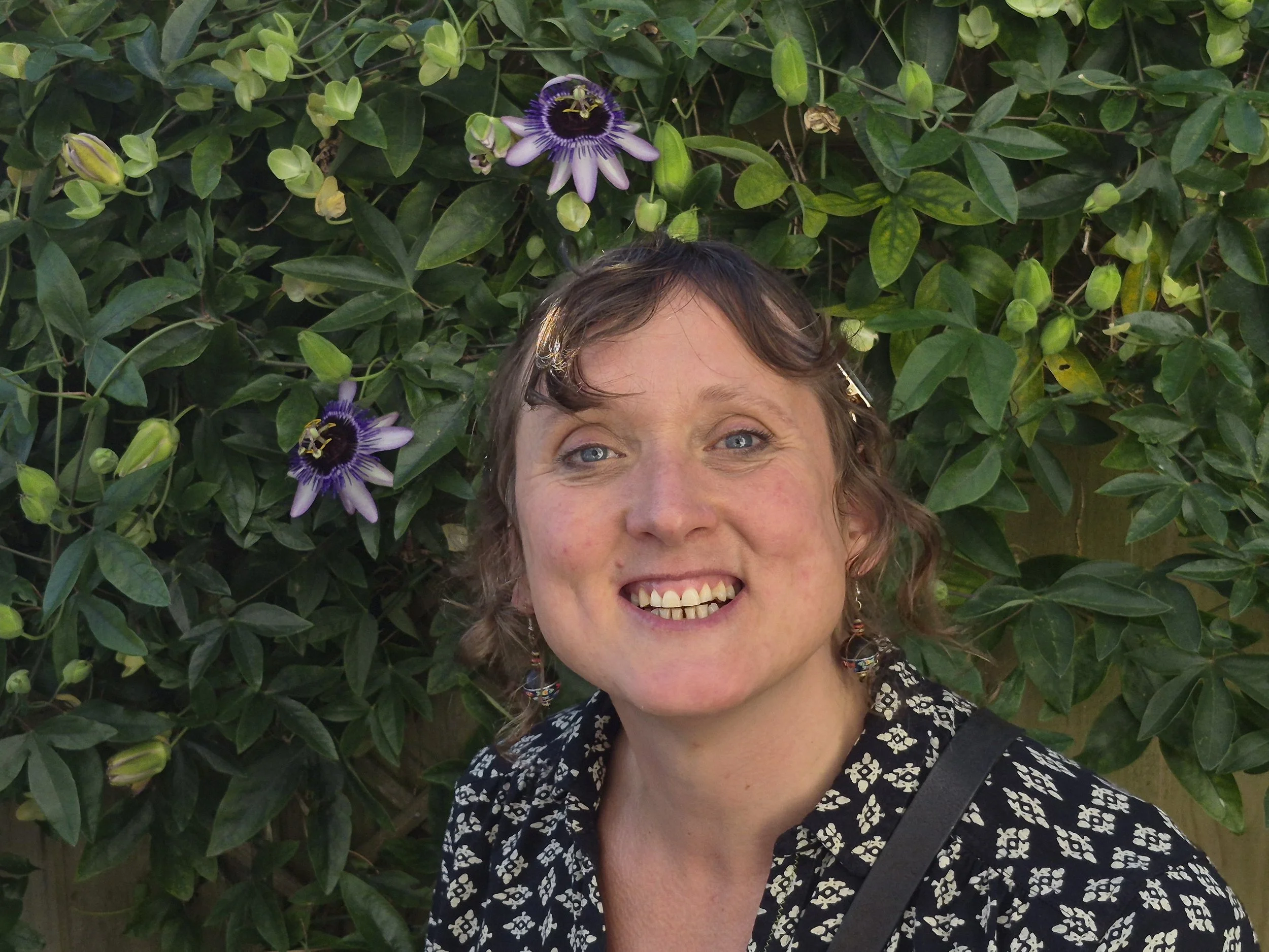 Jenny Wellwood is a white 46 year old UK woman, She has curly light brown hair and blue eyes, and a big smile- she is outdoors, surrounded by green leaves and purple passionflower blooming in the background.