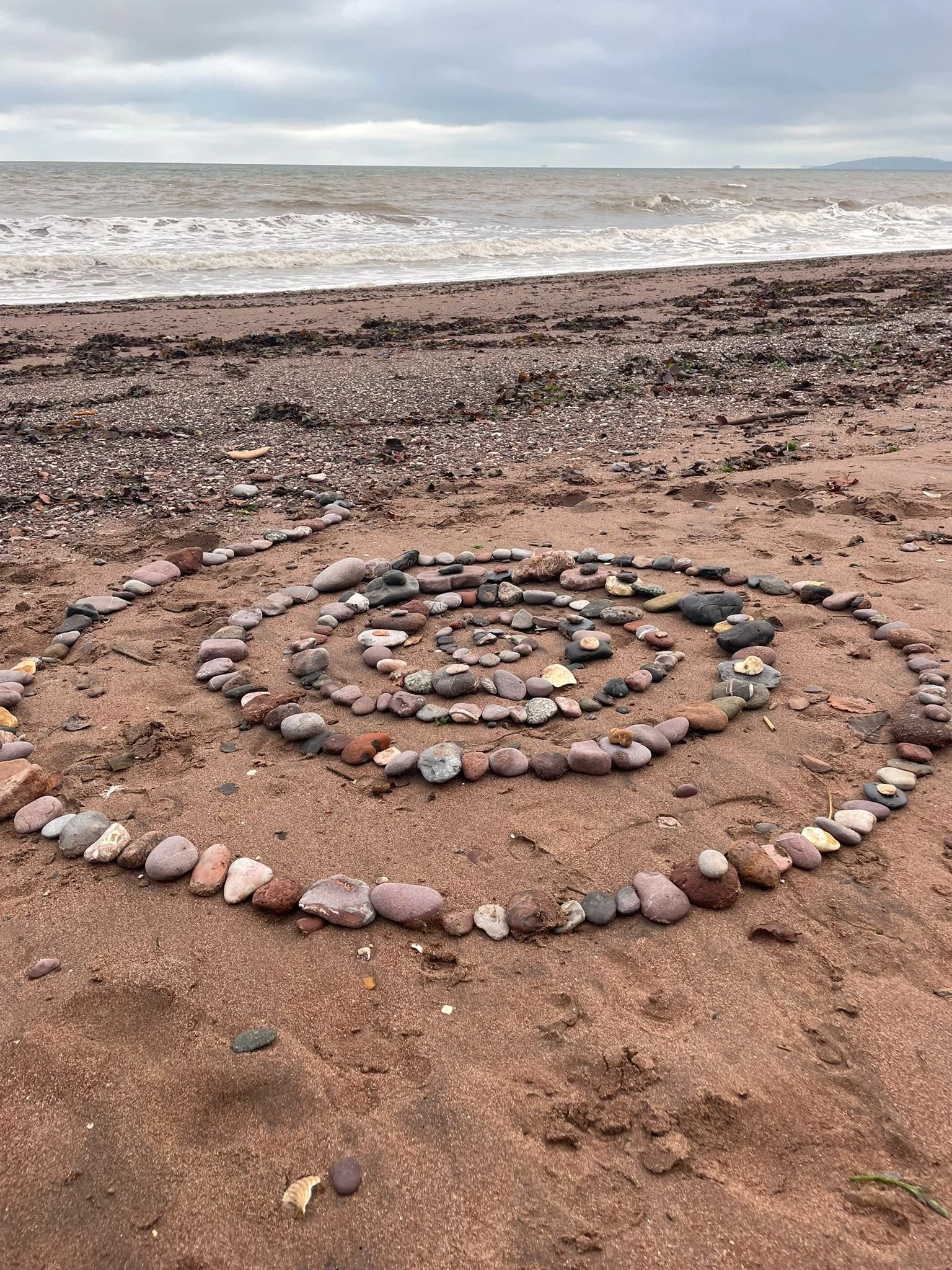 A spiral shape made from small and medium-sized smooth stones and pebbles on a sandy beach, with the ocean and cloudy sky in the background.