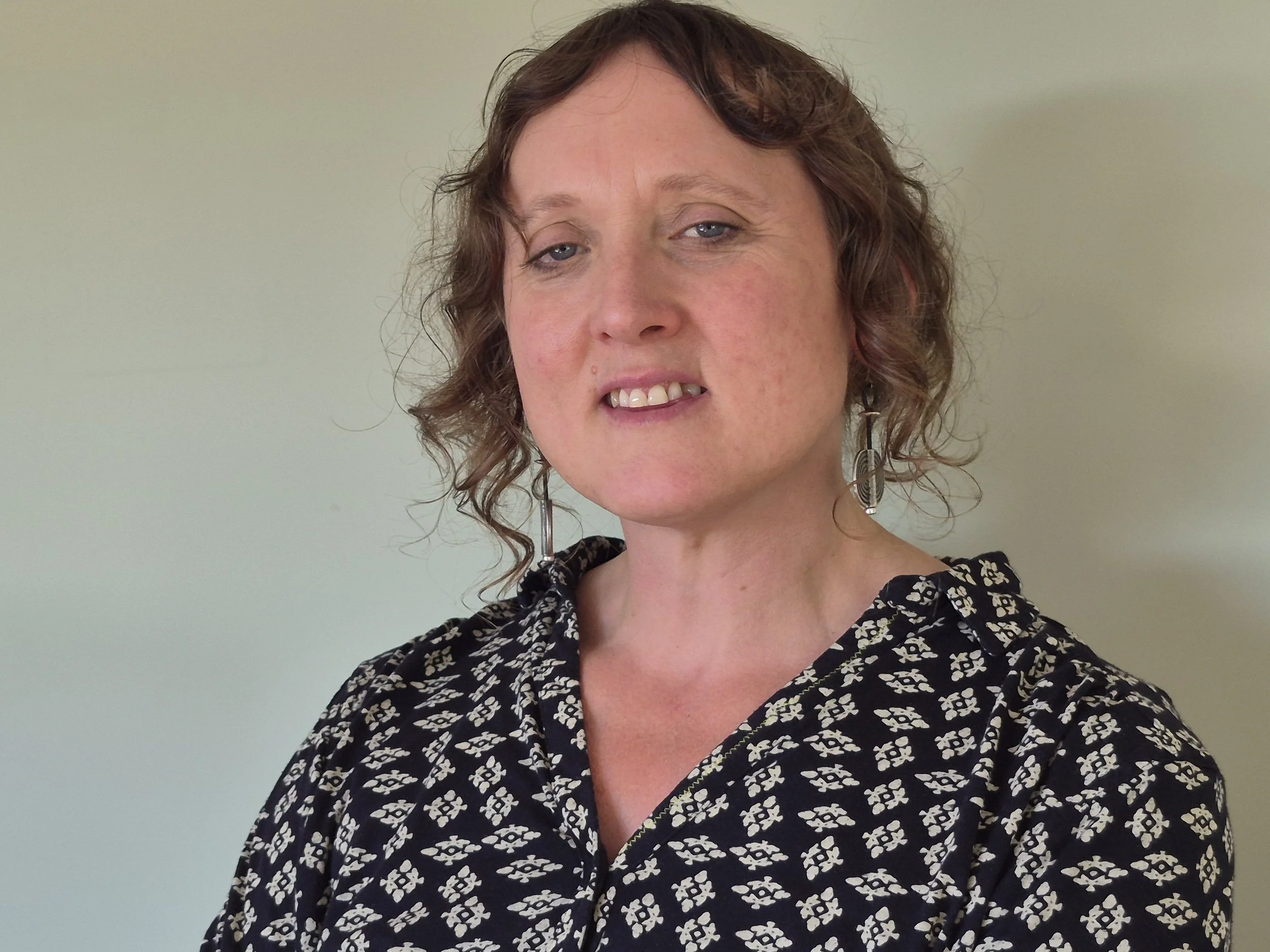 Jenny Wellwood is a 46 years old white woman, she has curly brown hair tied back, and is wearing a black and white patterned top. She is standing in front of a plain light green wall. She has a thoughtful posture and kind eyes.