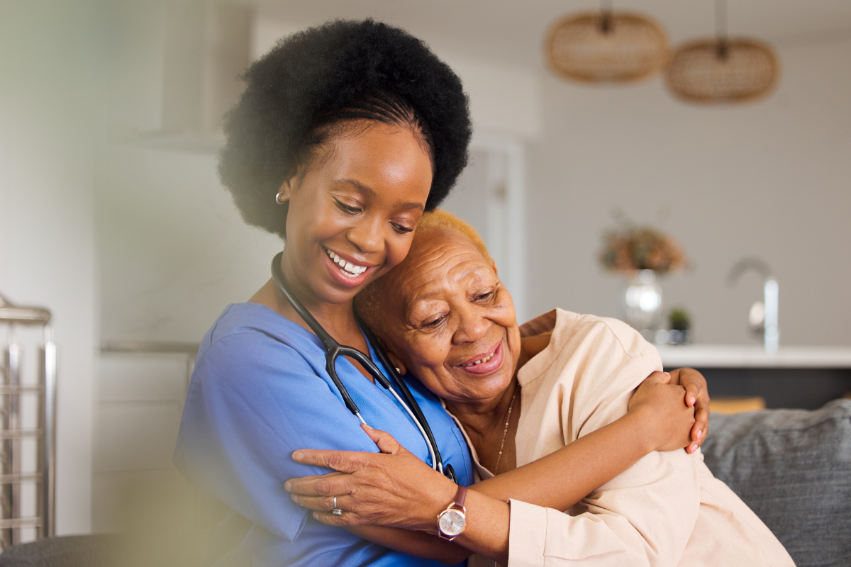 A young female healthcare worker in blue scrubs hugging an elderly woman with light brown skin and short blonde hair in a cozy home setting, both smiling.