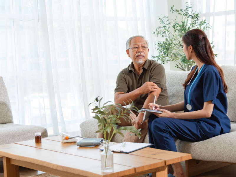 A female nurse or healthcare worker talking to an elderly man during a medical consultation in a bright room with plants and large windows.
