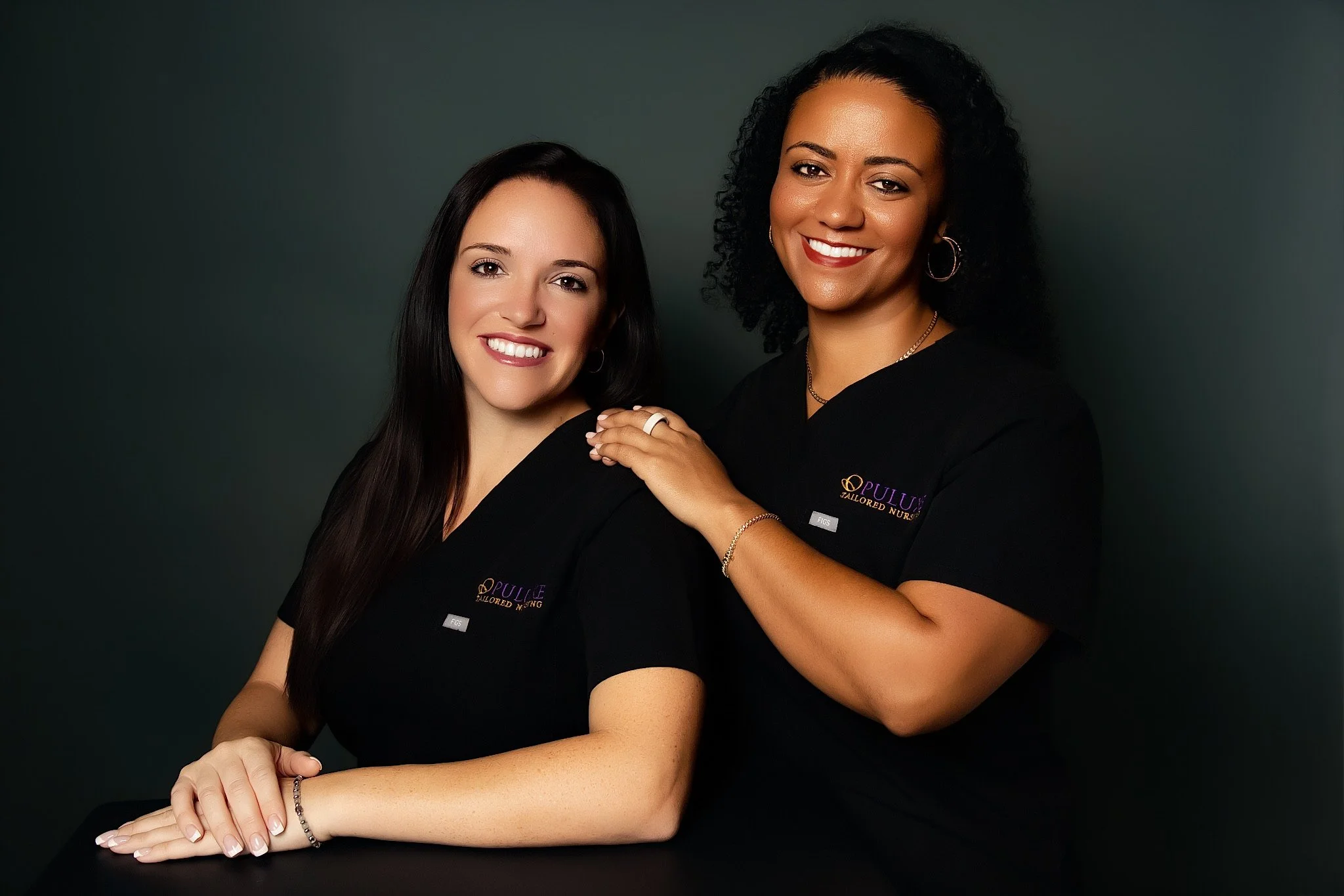 Two women, one with straight brown hair and one with curly black hair, smiling while dressed in black shirts with a logo for 'Pulley Tailored Nursing,' posing against a dark green background.