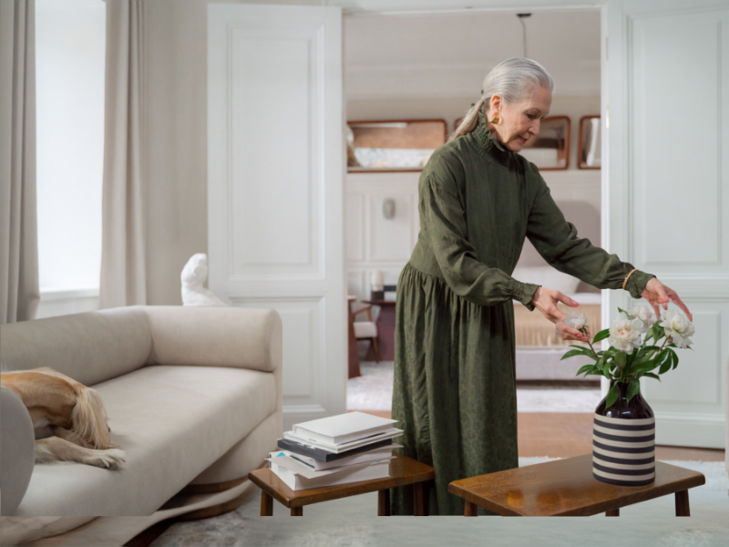An elderly woman with gray hair in a green dress arranging white flowers in a striped vase on a wooden table in a living room