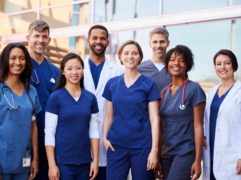Group of diverse healthcare professionals standing together outside a medical facility, smiling at the camera.