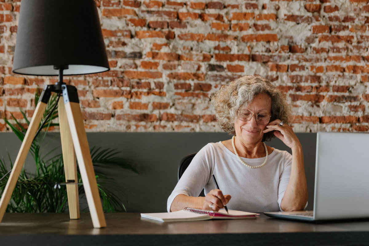 An elderly woman with curly gray hair, wearing glasses and a pearl necklace, sits at a desk in front of a laptop, writing in a notepad, with a smile.