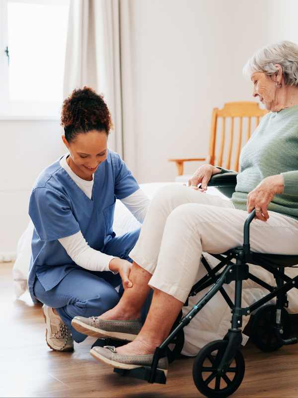 A nurse assisting an elderly woman in a wheelchair with her shoe in a well-lit room.
