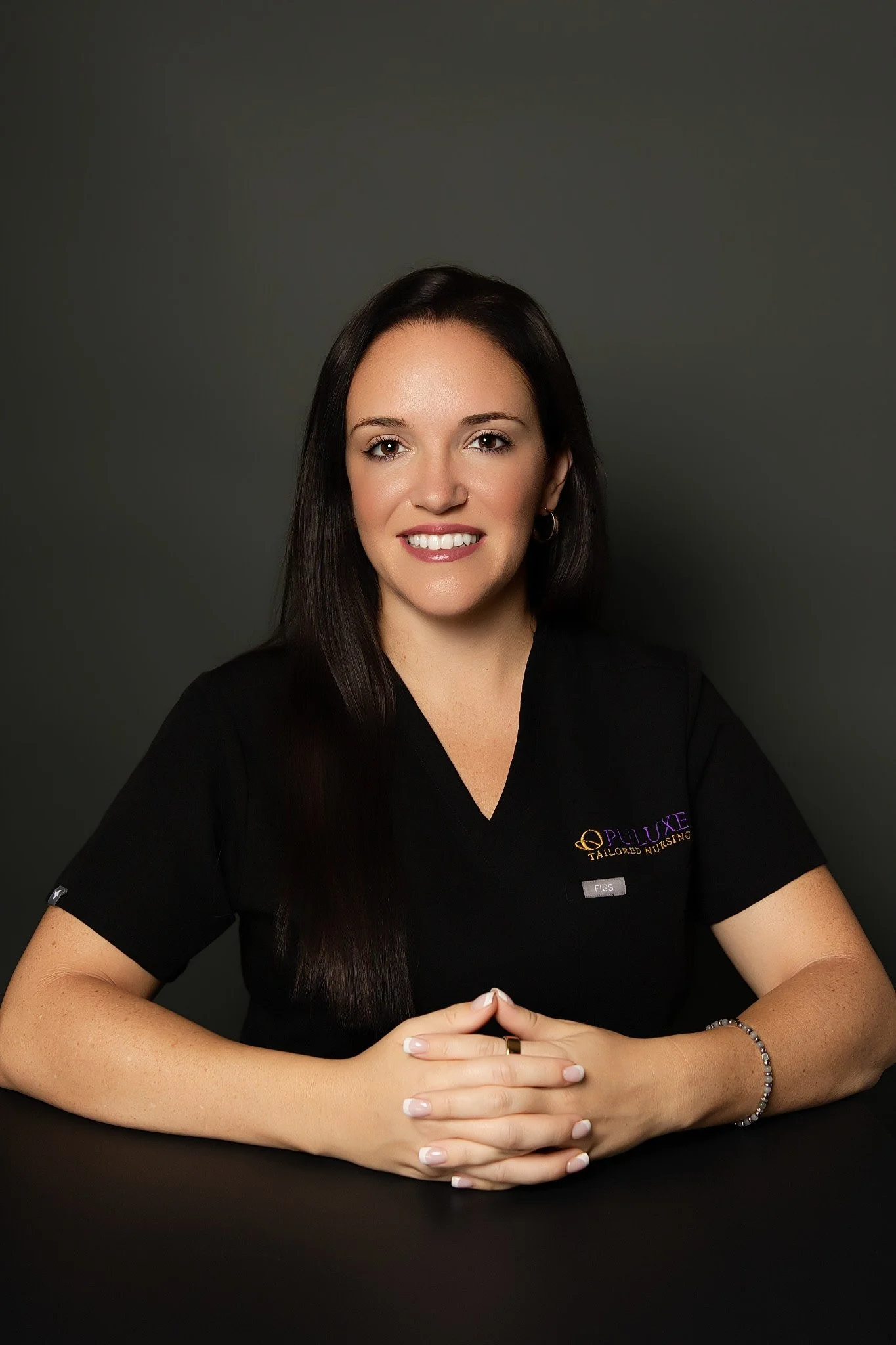 Portrait of a smiling woman with long dark hair, wearing a black medical uniform with a name badge, sitting at a black table with hands clasped.