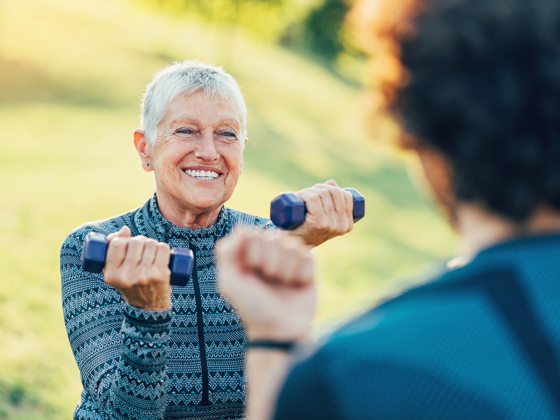 A smiling elderly woman exercising outdoors, lifting small blue dumbbells in each hand.