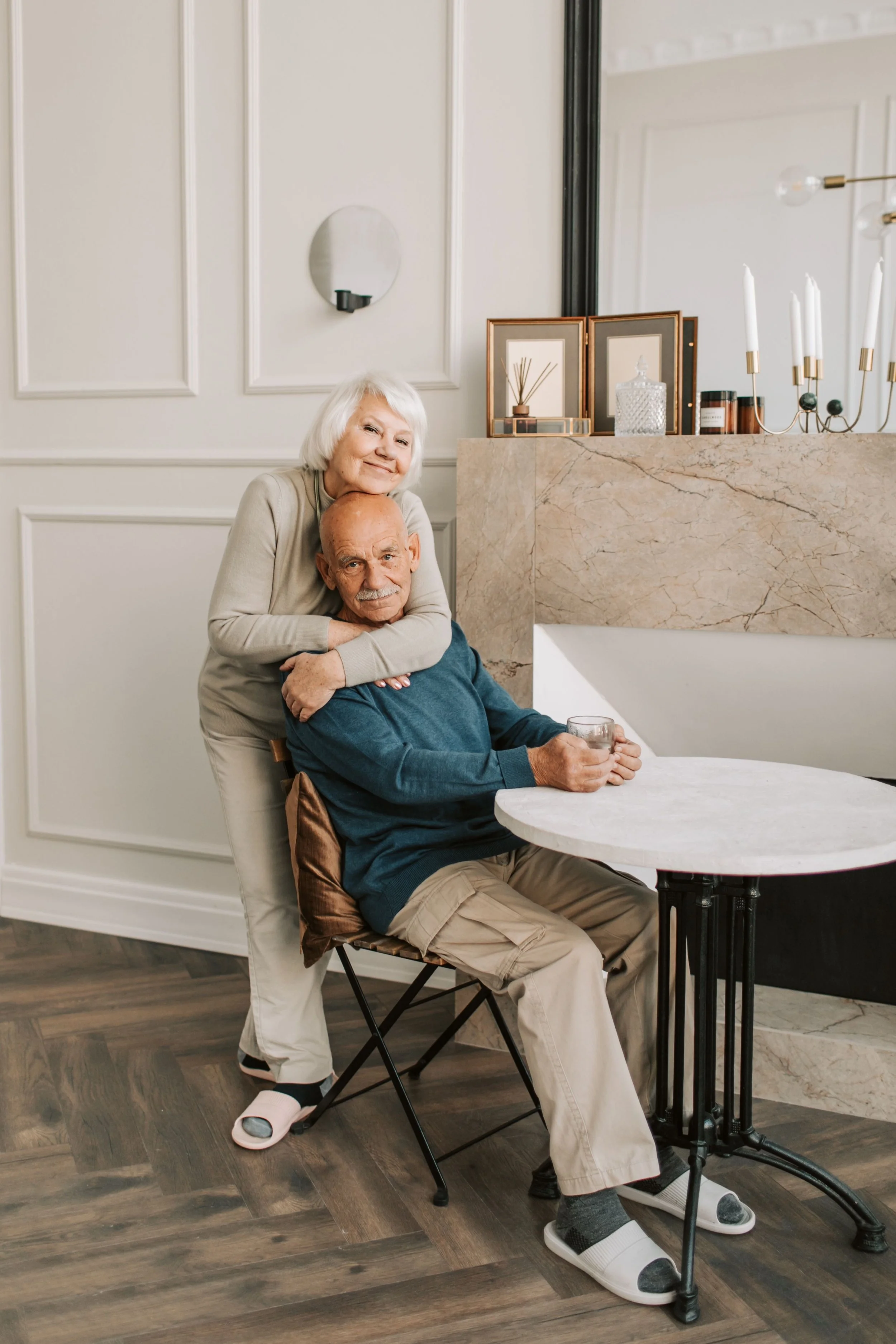 An elderly woman with white hair hugging an elderly man with a bald head and gray beard in a cozy, elegant room. The woman is smiling and resting her head on the man's shoulder while the man, seated at a table, holds a glass of water. The room has neutral-colored walls, decorative framed artwork, a marble mantel, and a large mirror. The floor is wood, and the man is wearing beige pants, gray socks, and slippers, with a blue sweater.